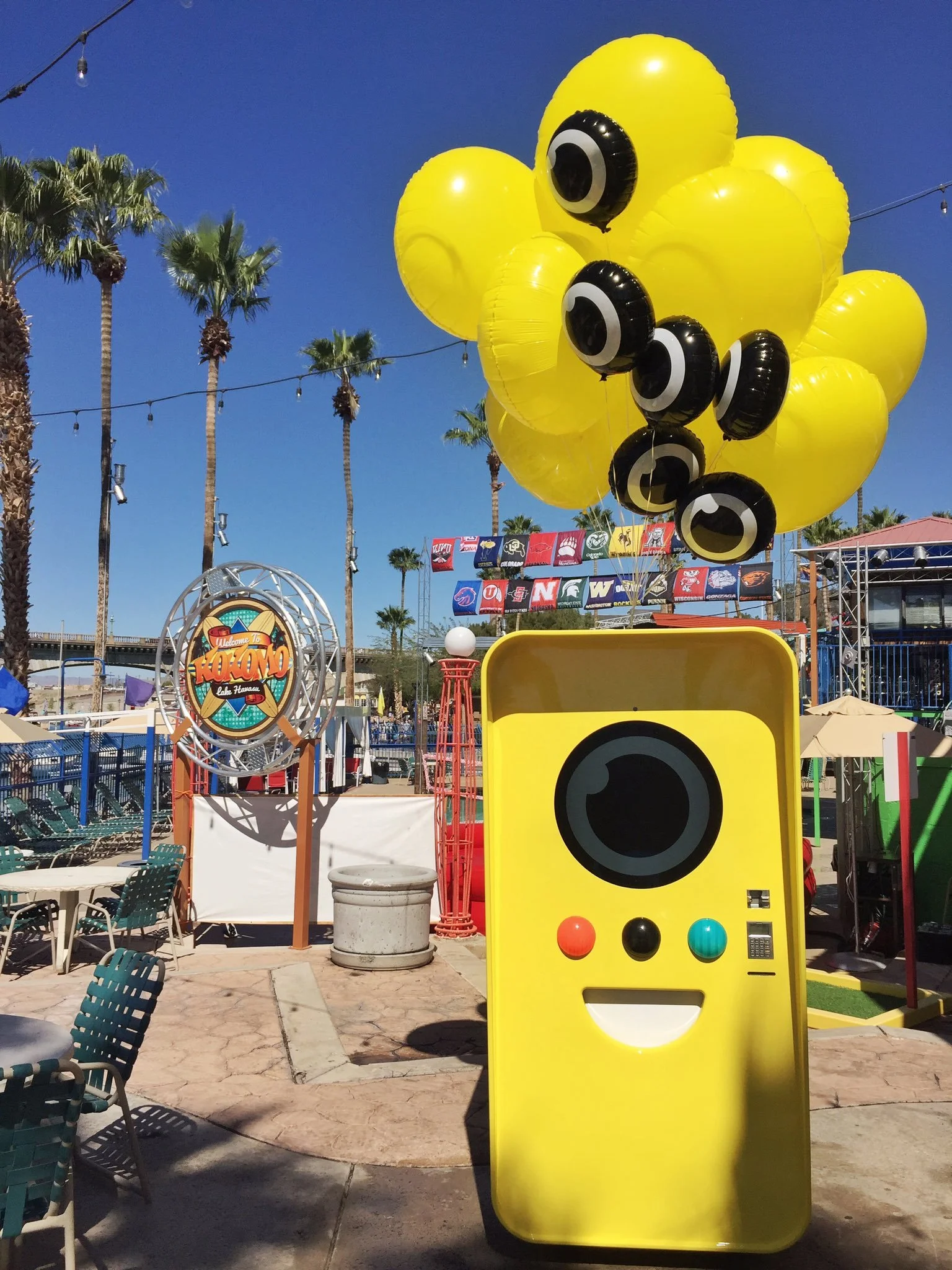 Yellow Snapbot vending machine with a big camera-lens eye and balloons at an outdoor patio, Welcome to Kokomo sign behind