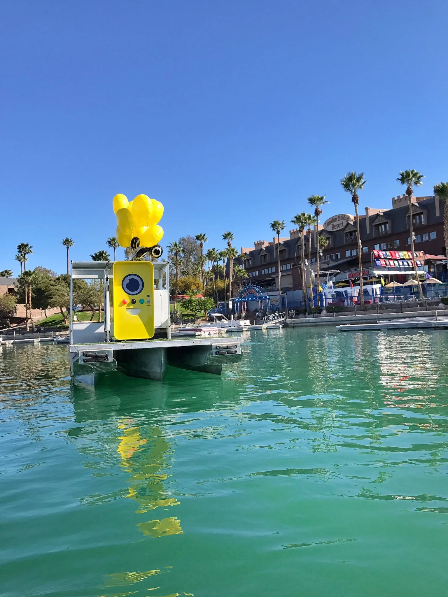 Yellow Snapbot vending machine with balloons on a floating dock in green water, London Bridge Resort behind