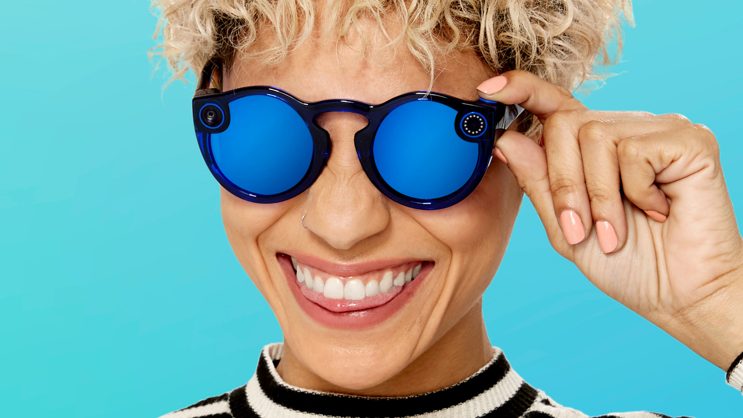 Smiling person with curly blonde hair wearing blue Snap Spectacles with mirrored lenses on a teal background