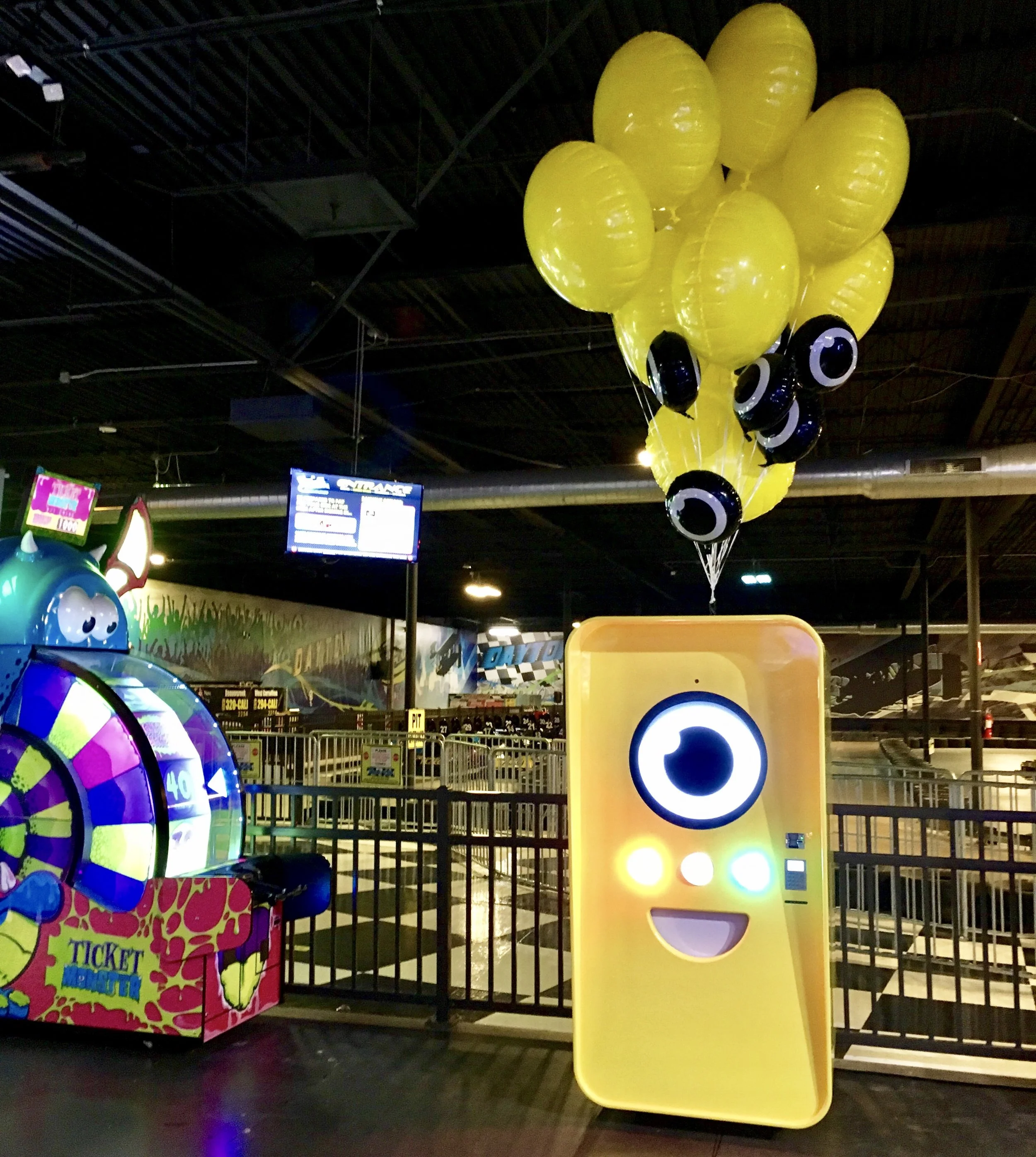 Yellow Snapbot vending machine with lit face and balloons by a go-kart track and Ticket Monster arcade game.
