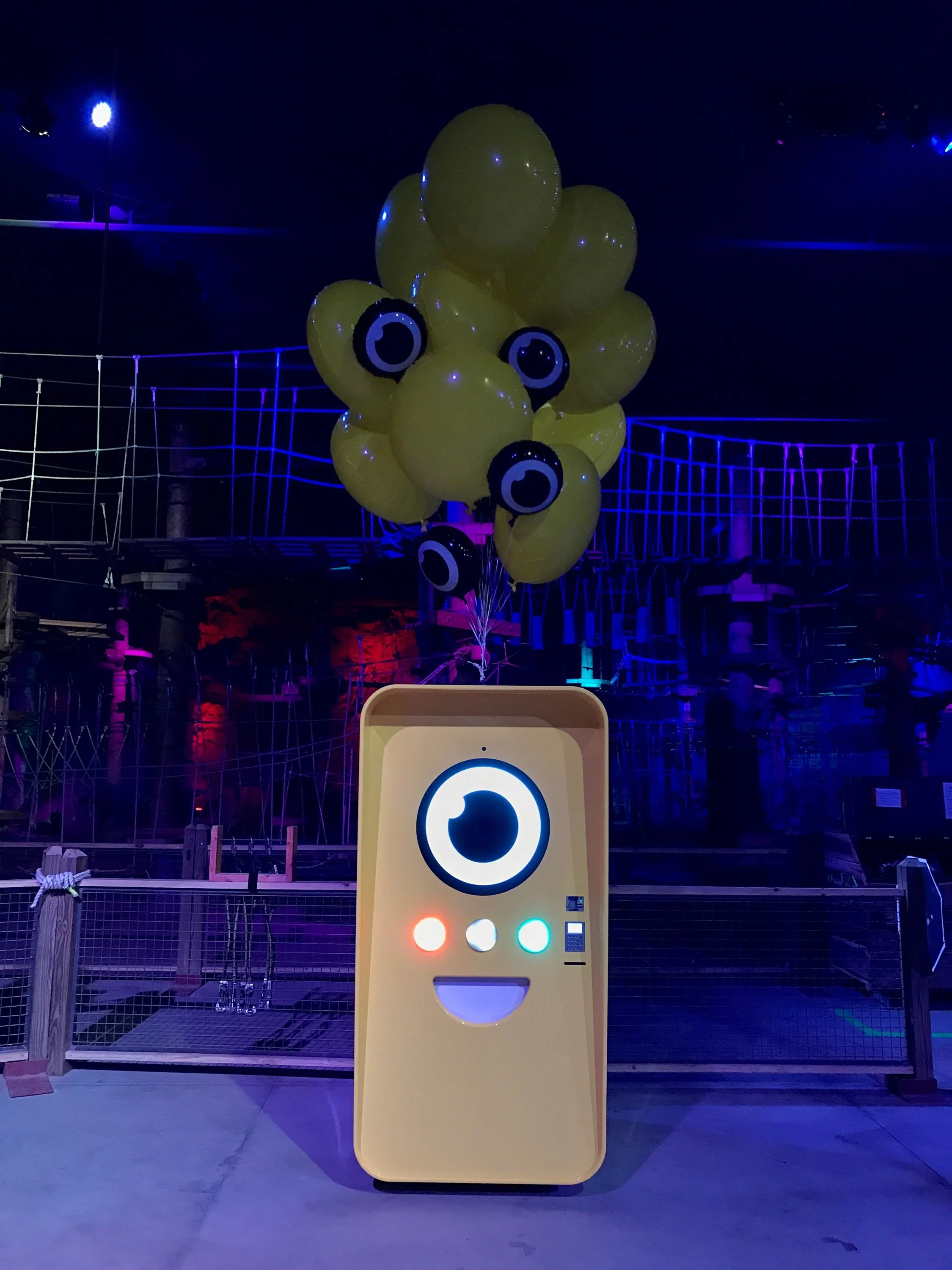Cream-yellow Snapbot vending machine with lit eye and balloons in a dark ropes course lit blue and purple.