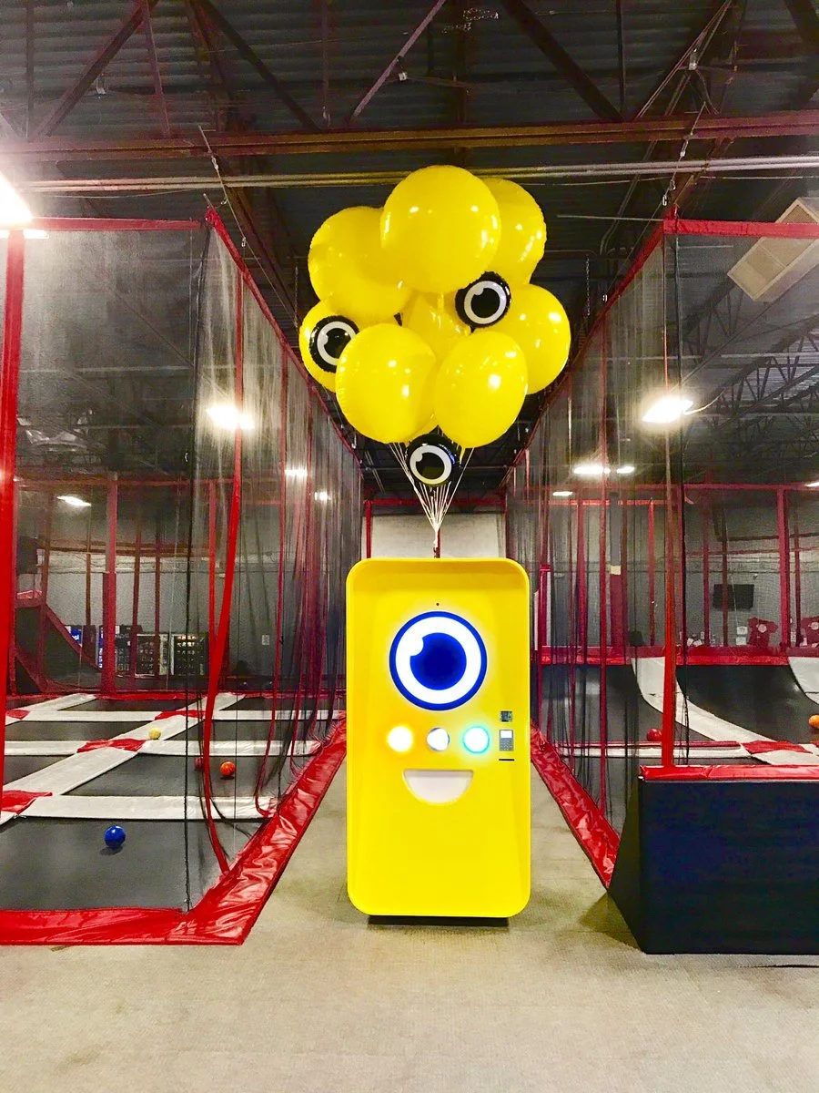Yellow Snapbot vending machine with balloons on a trampoline court framed by red-padded netting.