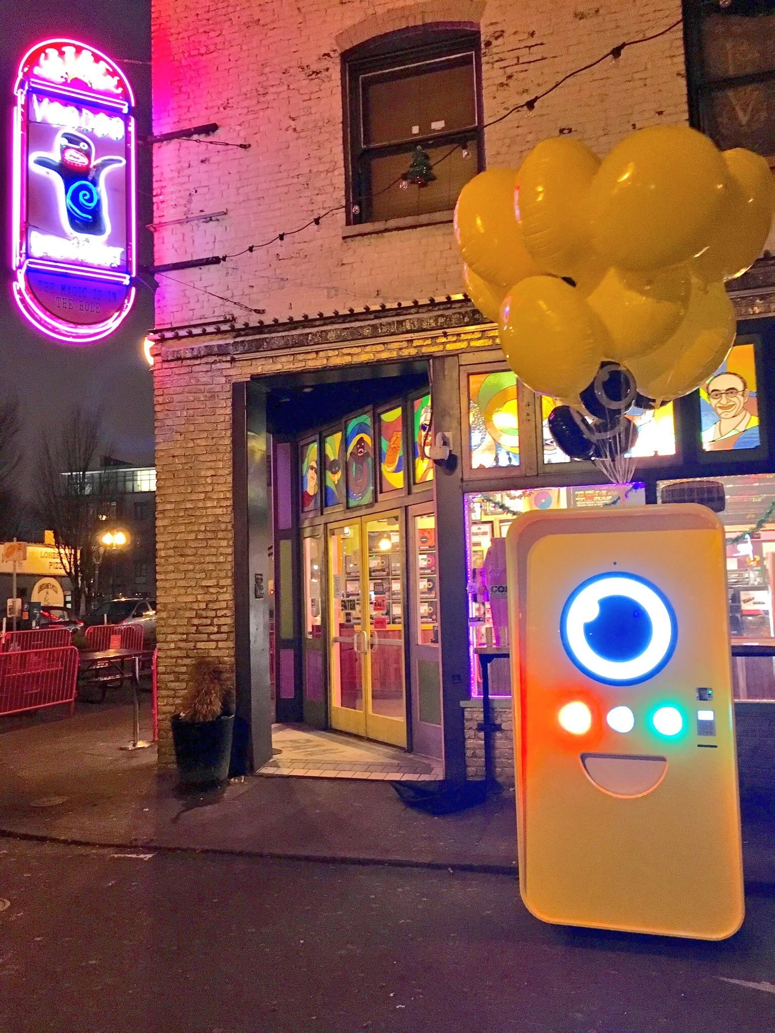 Yellow Snapbot Spectacles vending machine with glowing face and balloons at night outside a brick building with a neon penguin sign.