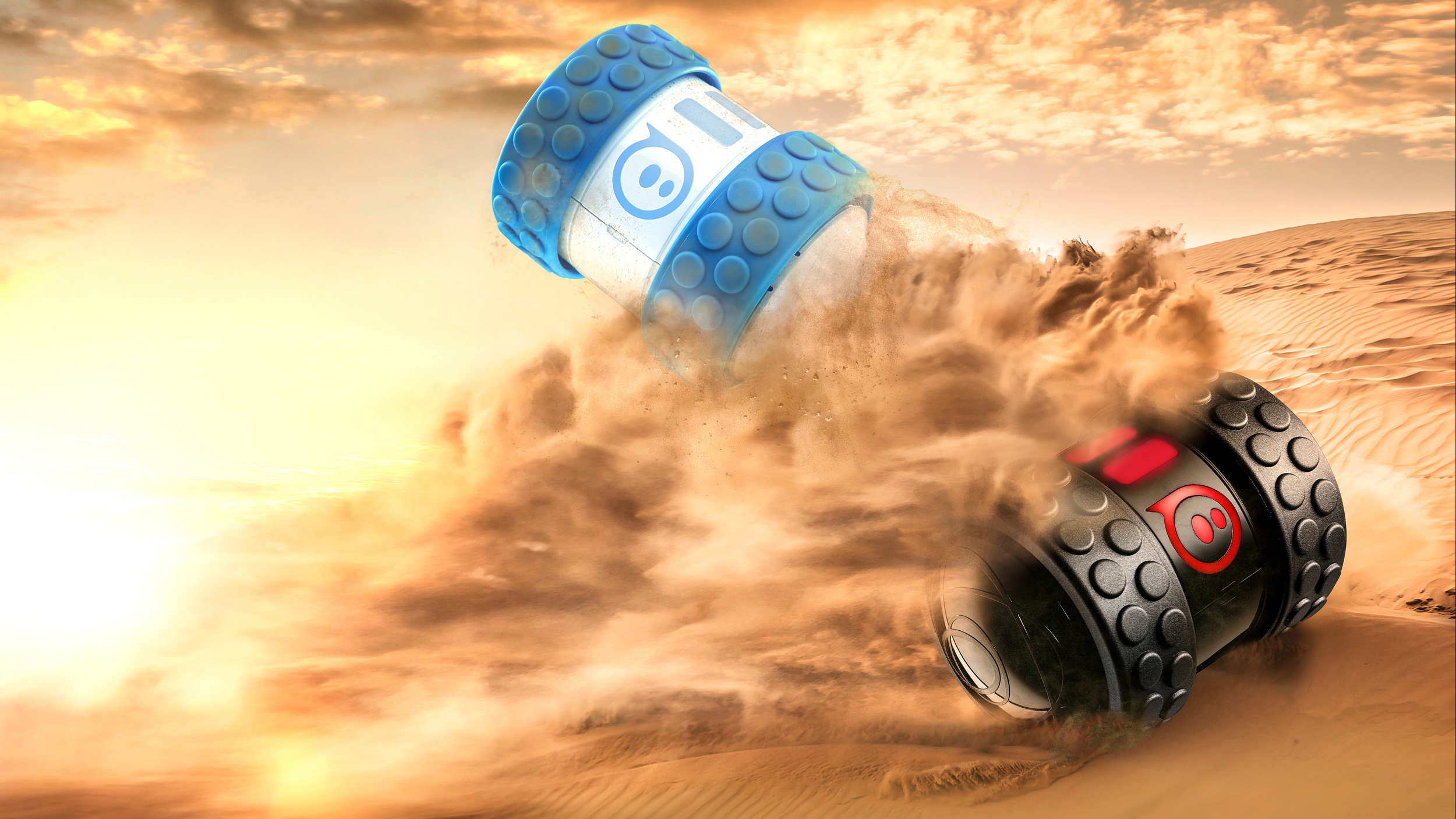 Blue and black Sphero Ollie robots speeding across desert sand dunes at sunset, throwing up dust