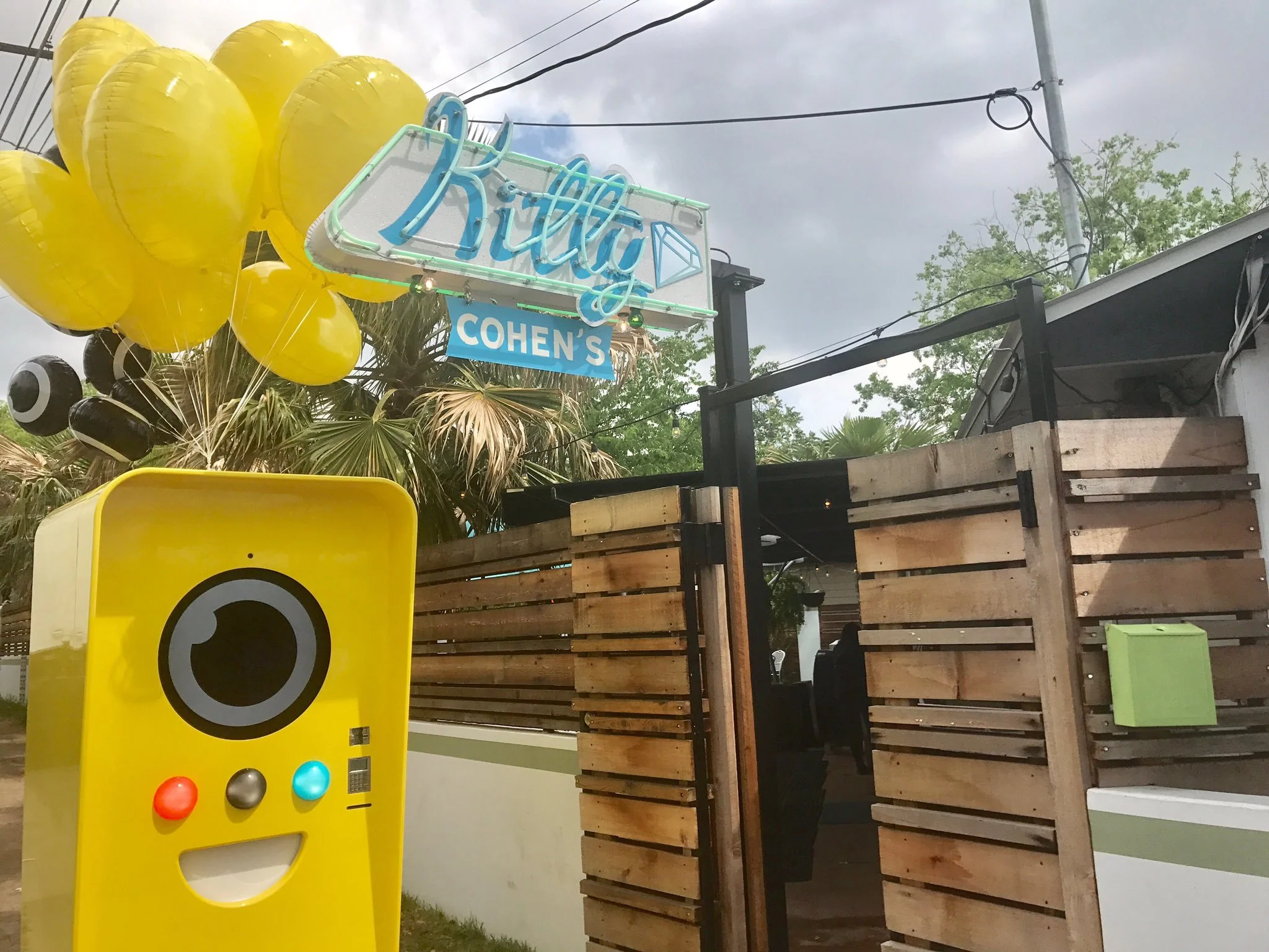 Yellow Snapbot machine with balloons under a neon Kitty Cohen's sign and wooden fence