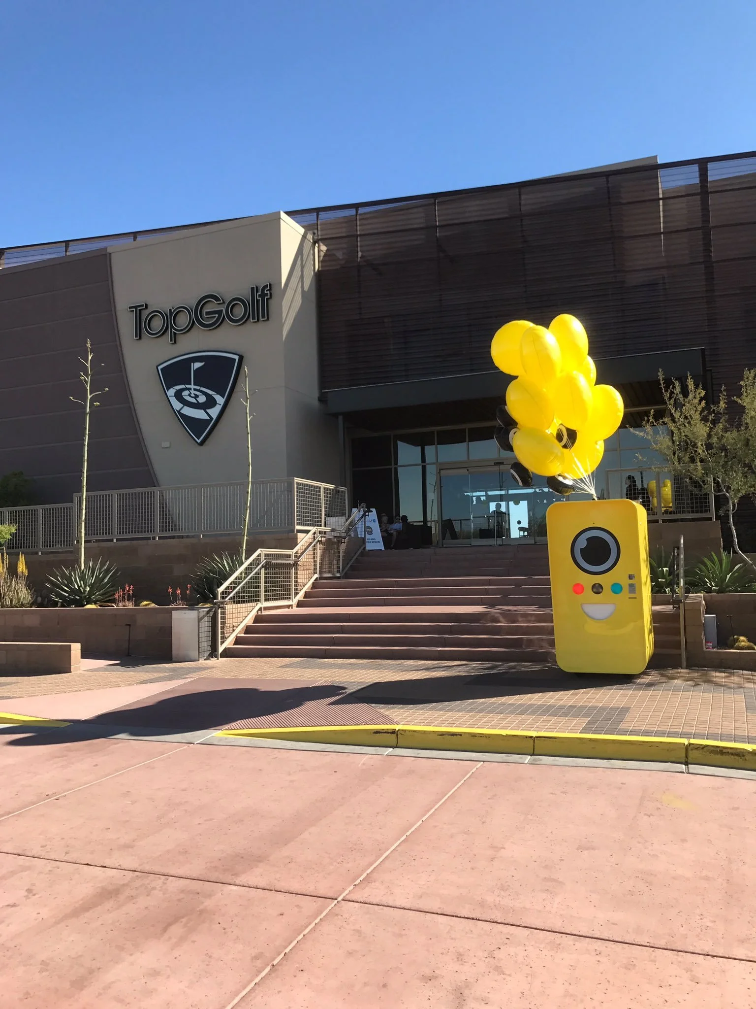 Yellow Snapbot vending machine with balloons at the foot of steps below a TopGolf building sign