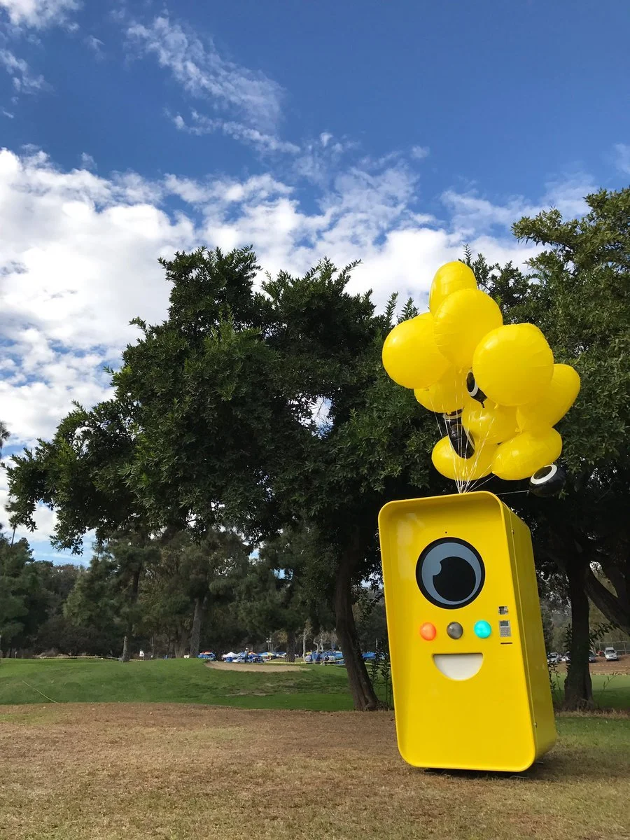 Yellow Snapbot Spectacles vending machine with a camera-lens face and yellow balloons on a grassy park lawn under blue sky.