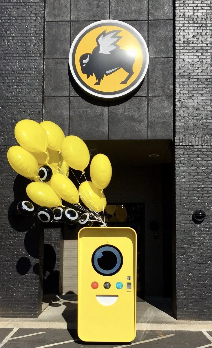 Yellow Snapbot vending machine with balloons at a black-brick entrance below the Buffalo Wild Wings logo.