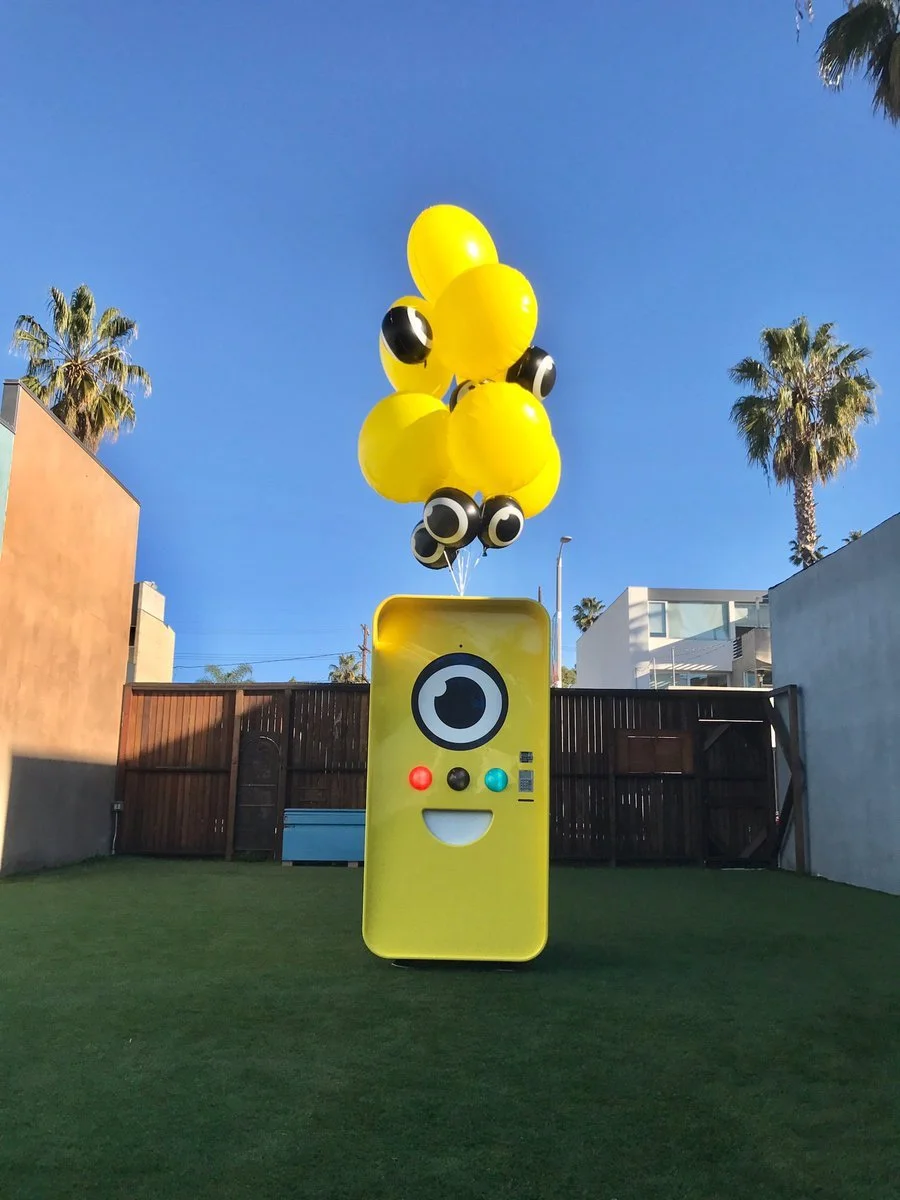 Yellow Snapbot vending machine with balloons on artificial turf, palm trees and clear blue sky behind.