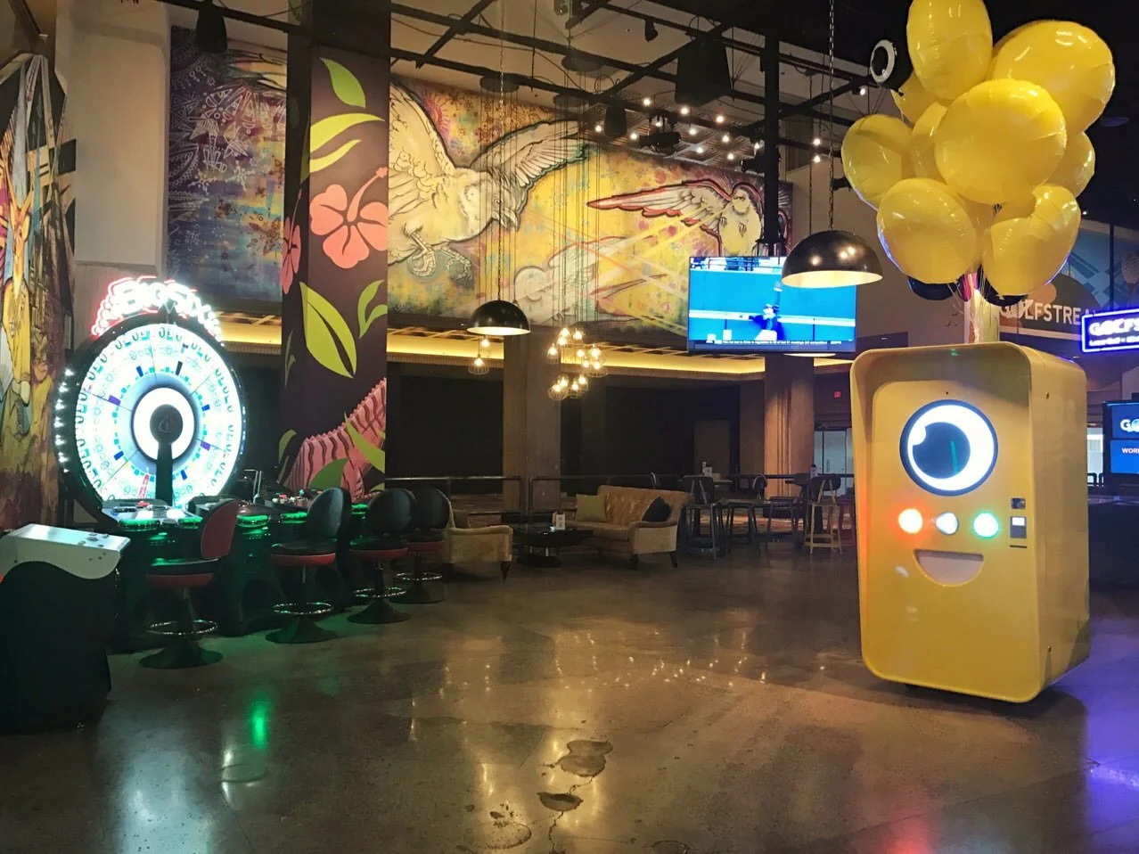 Yellow Snapbot vending machine with balloons inside a dim lounge with murals and a lit prize wheel