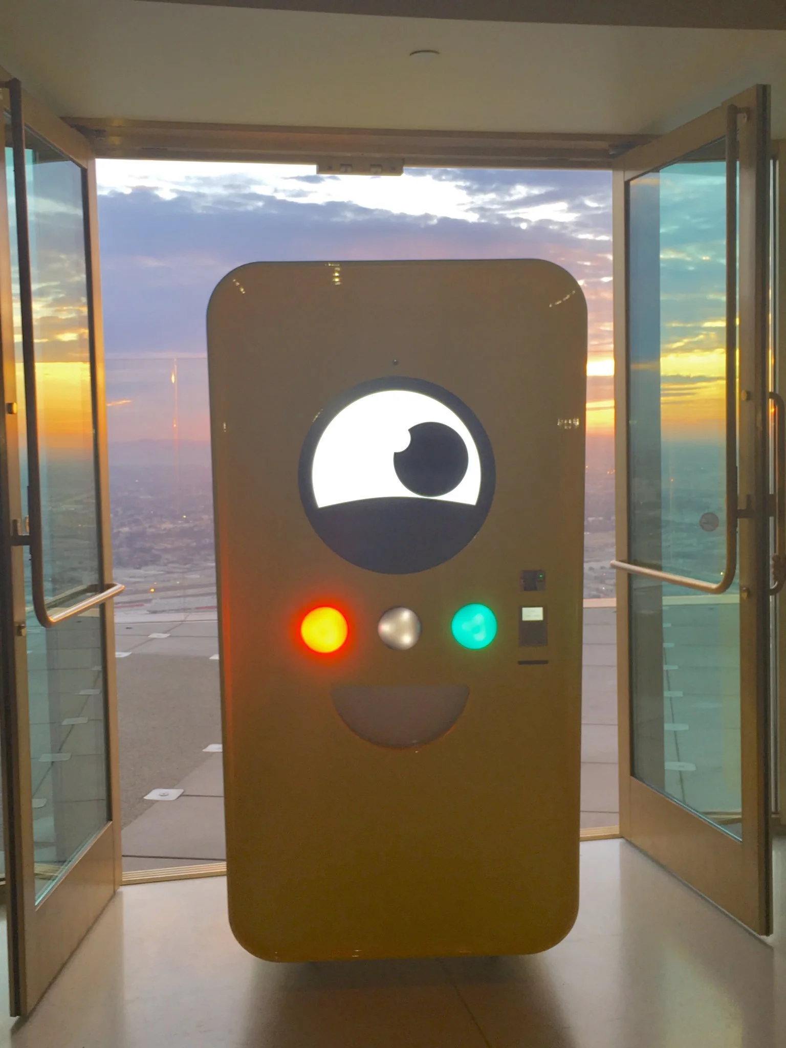 Yellow Snapbot Spectacles vending machine with a lit-up face, framed by open glass doors over a city skyline at sunset.