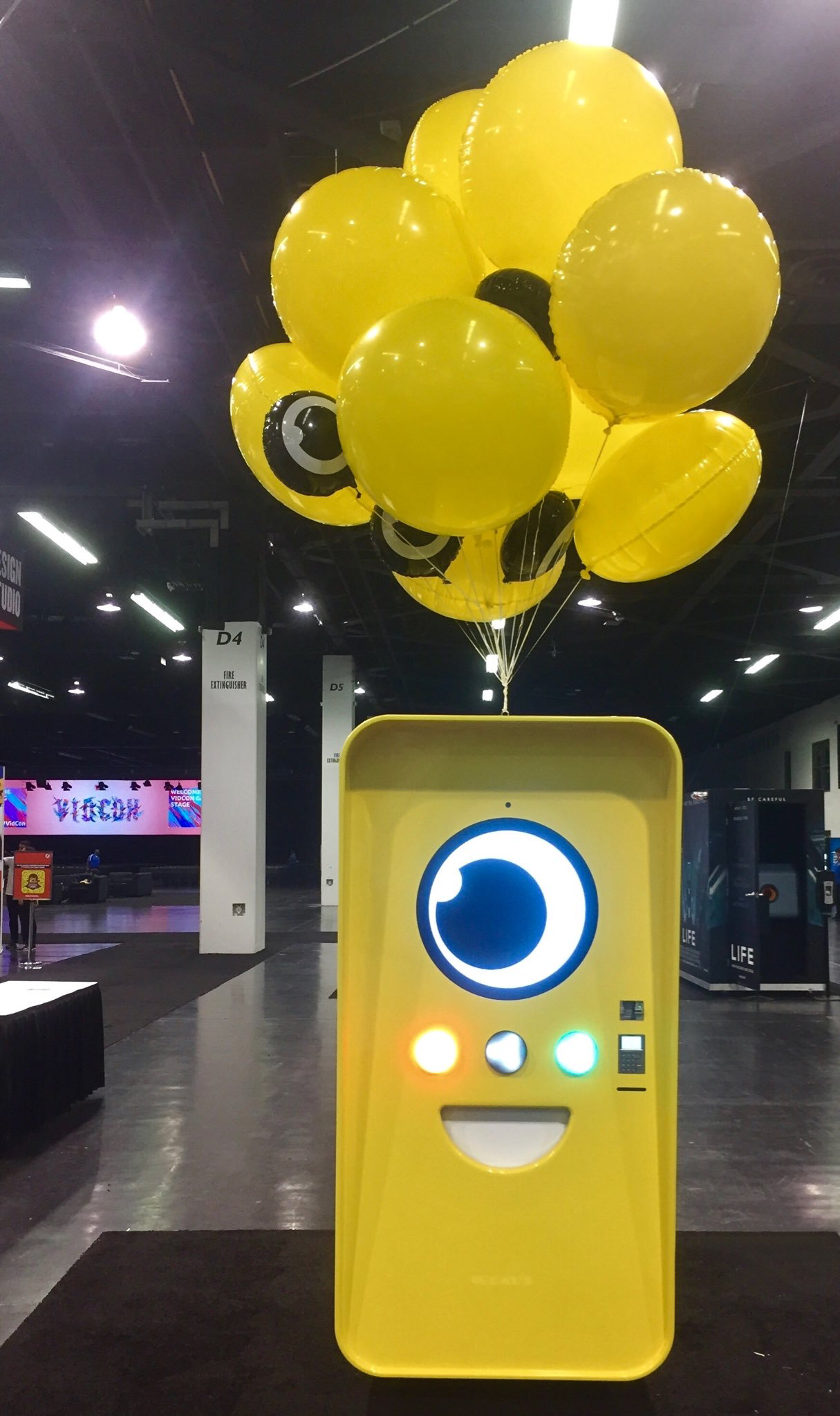 Yellow Snapbot Spectacles vending machine with balloons on a dark convention floor, VidCon screen in background