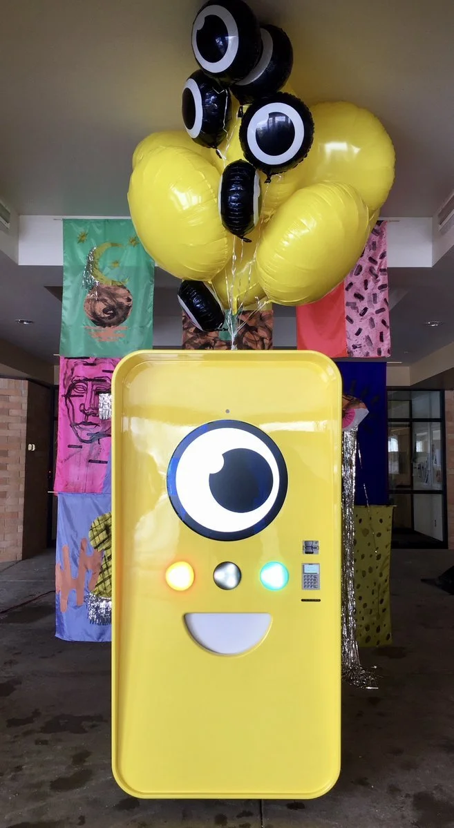 Yellow Snapbot vending machine and balloons indoors in front of colorful hand-painted hanging banners.