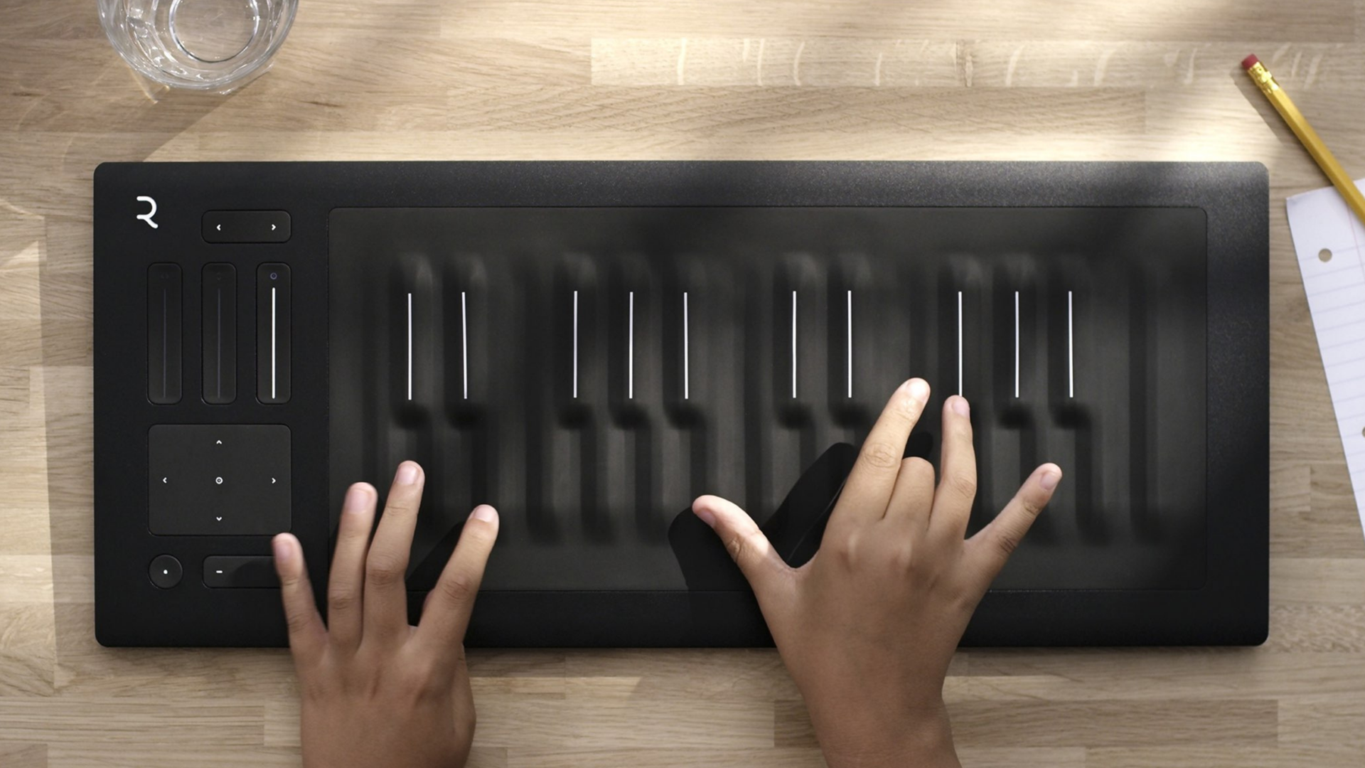 Top-down view of two hands playing a black ROLI Seaboard on a wooden desk beside a glass of water and pencil.