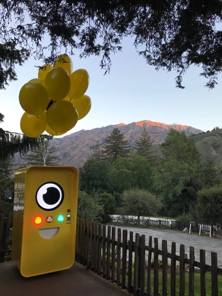 Yellow Snapchat Snapbot vending machine with a cartoon eye face and yellow balloons, on a path before Big Sur hills at dusk.
