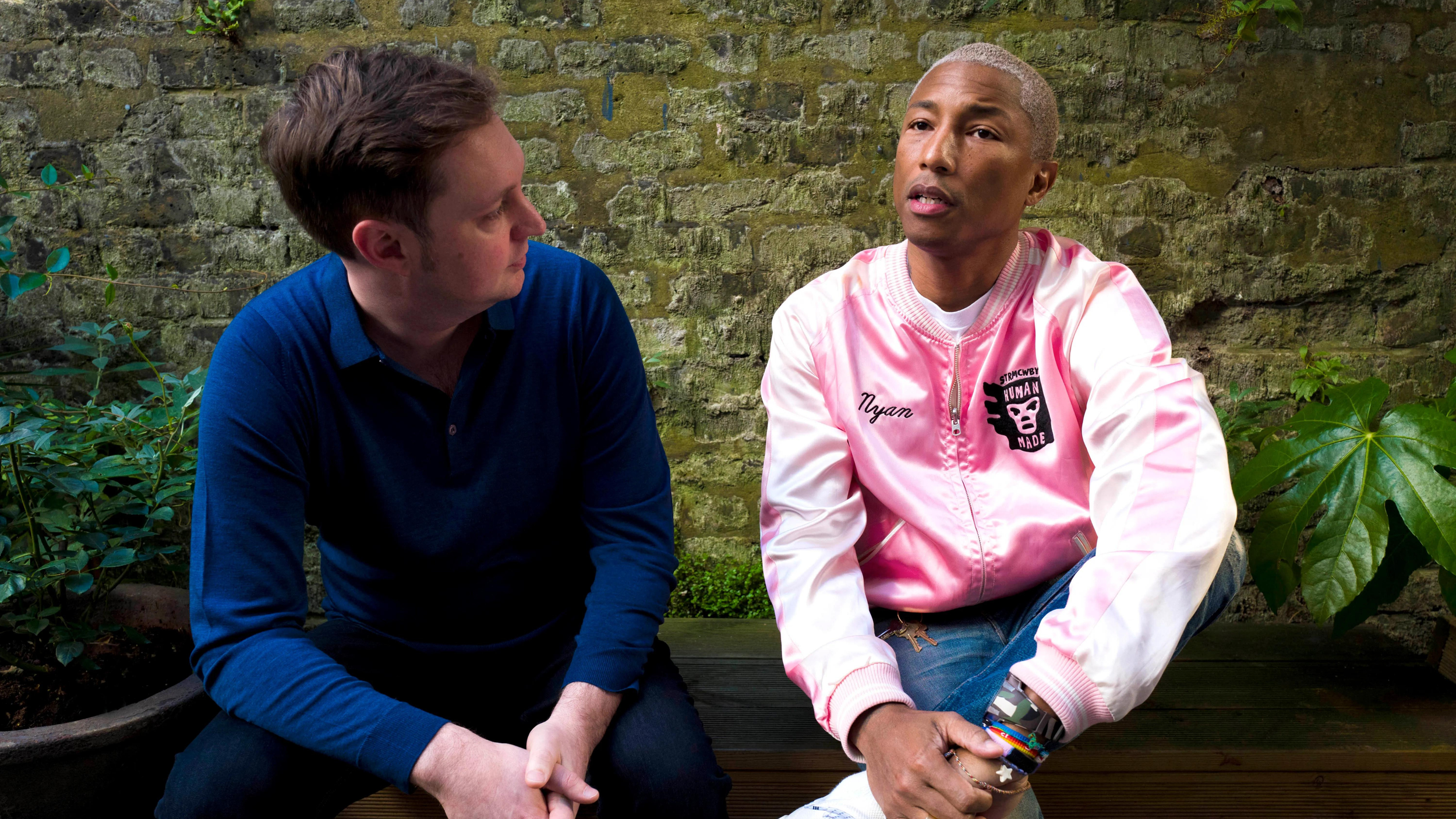 Two men seated on a bench by a mossy brick wall; one in a blue sweater, the other in a pink satin bomber jacket