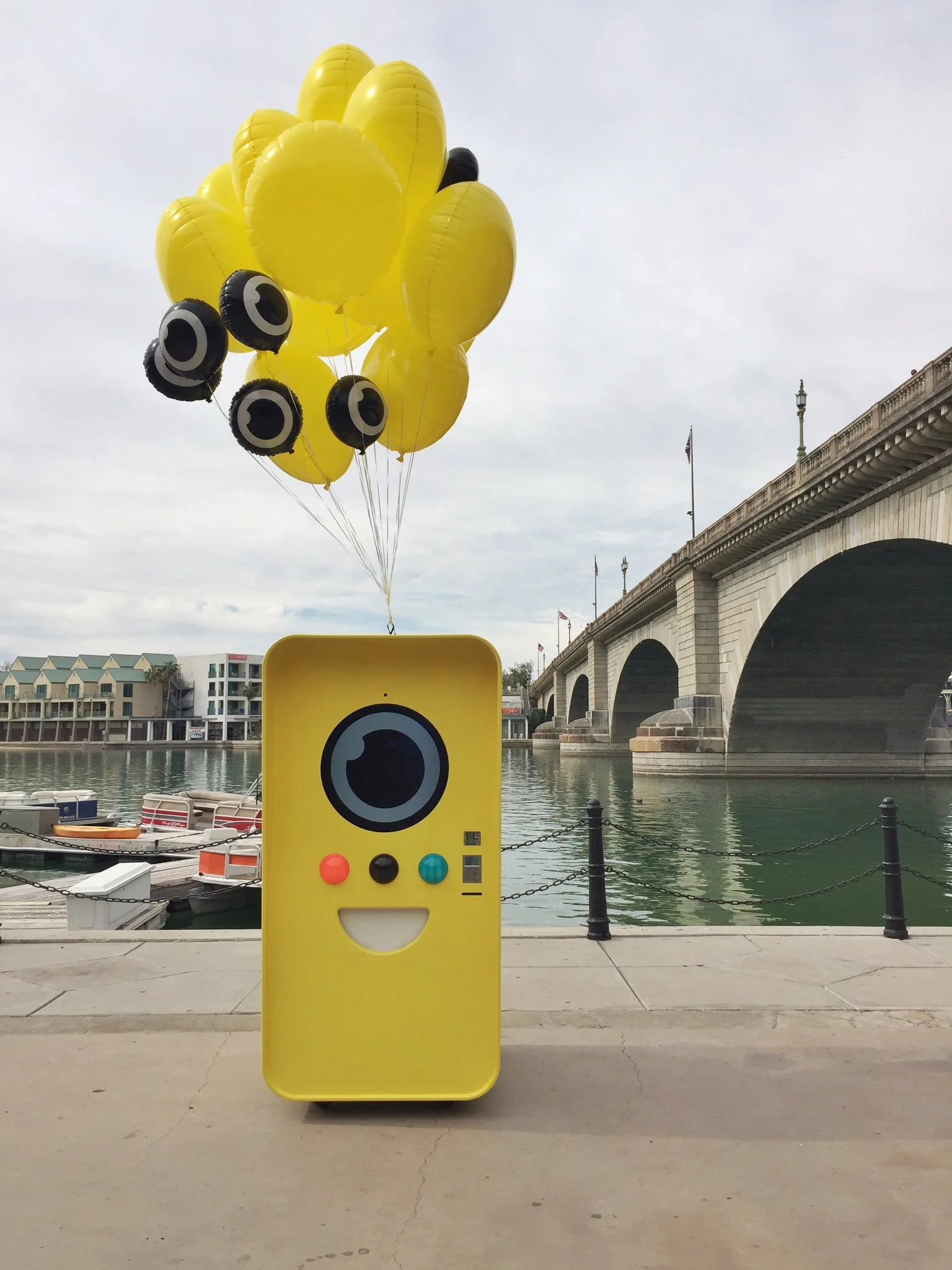 Yellow Snapbot vending machine with a smiling face and balloons on a walkway beside the stone London Bridge