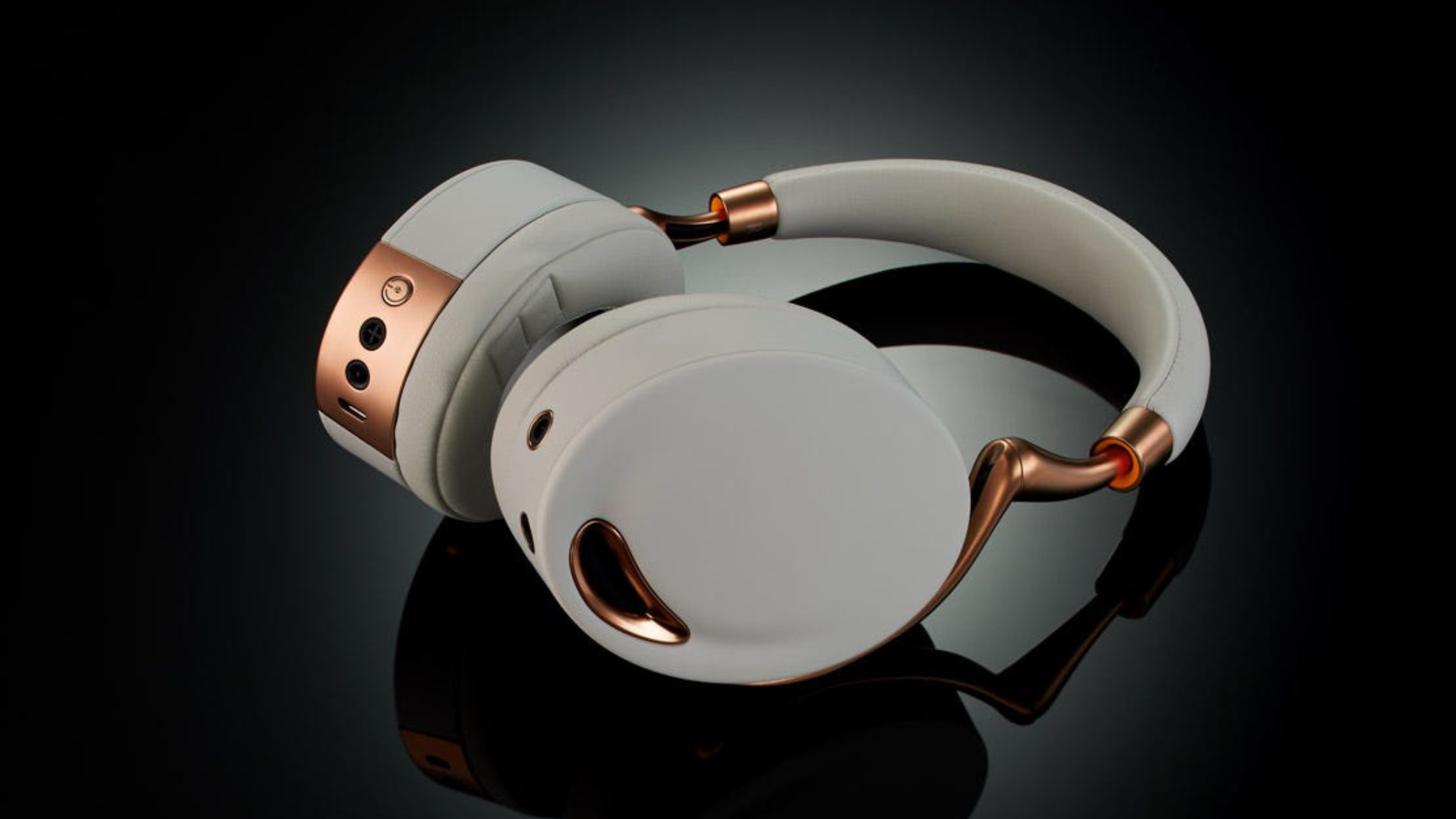 White and rose-gold over-ear headphone lying on a reflective black surface, showing control jacks.