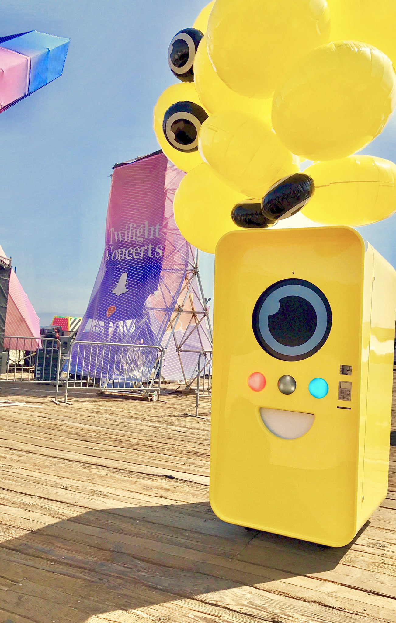 Yellow Snapbot Spectacles vending machine with balloons on a boardwalk beside a Twilight Concerts banner