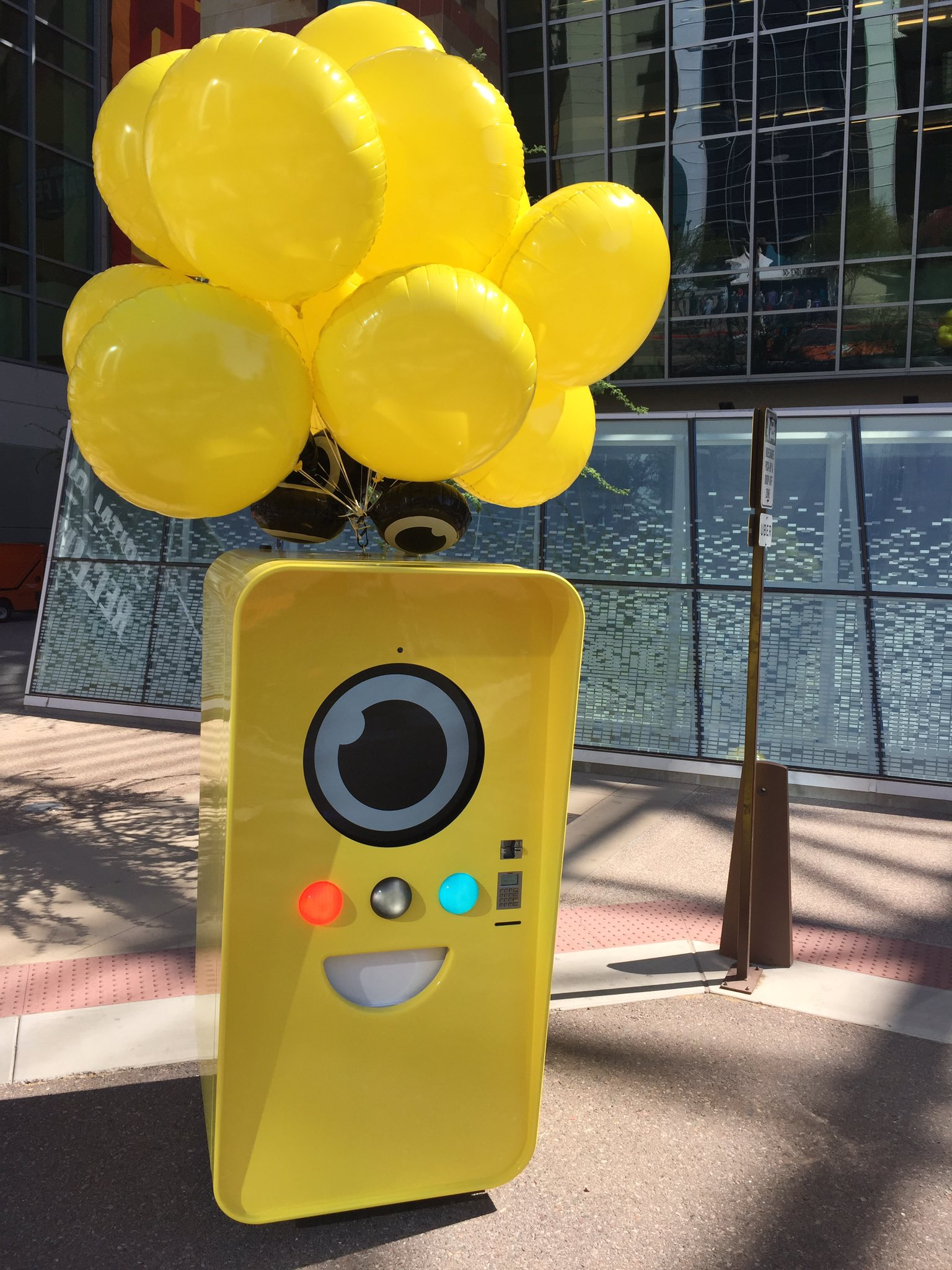 Snap Snapbot vending machine, a tall yellow box with a camera-lens face, topped with yellow balloons on a plaza
