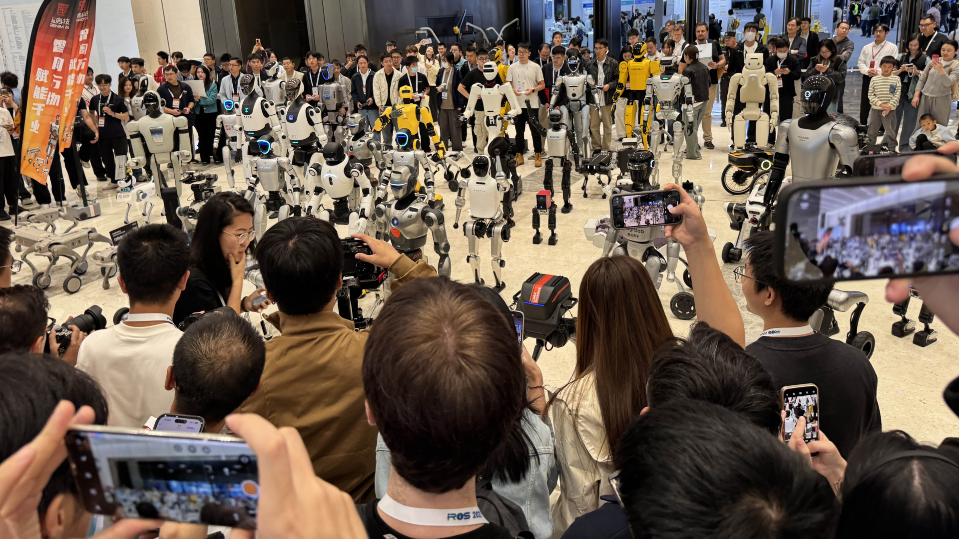 Crowd holding up phones to photograph a lineup of dozens of humanoid and quadruped robots in a convention hall