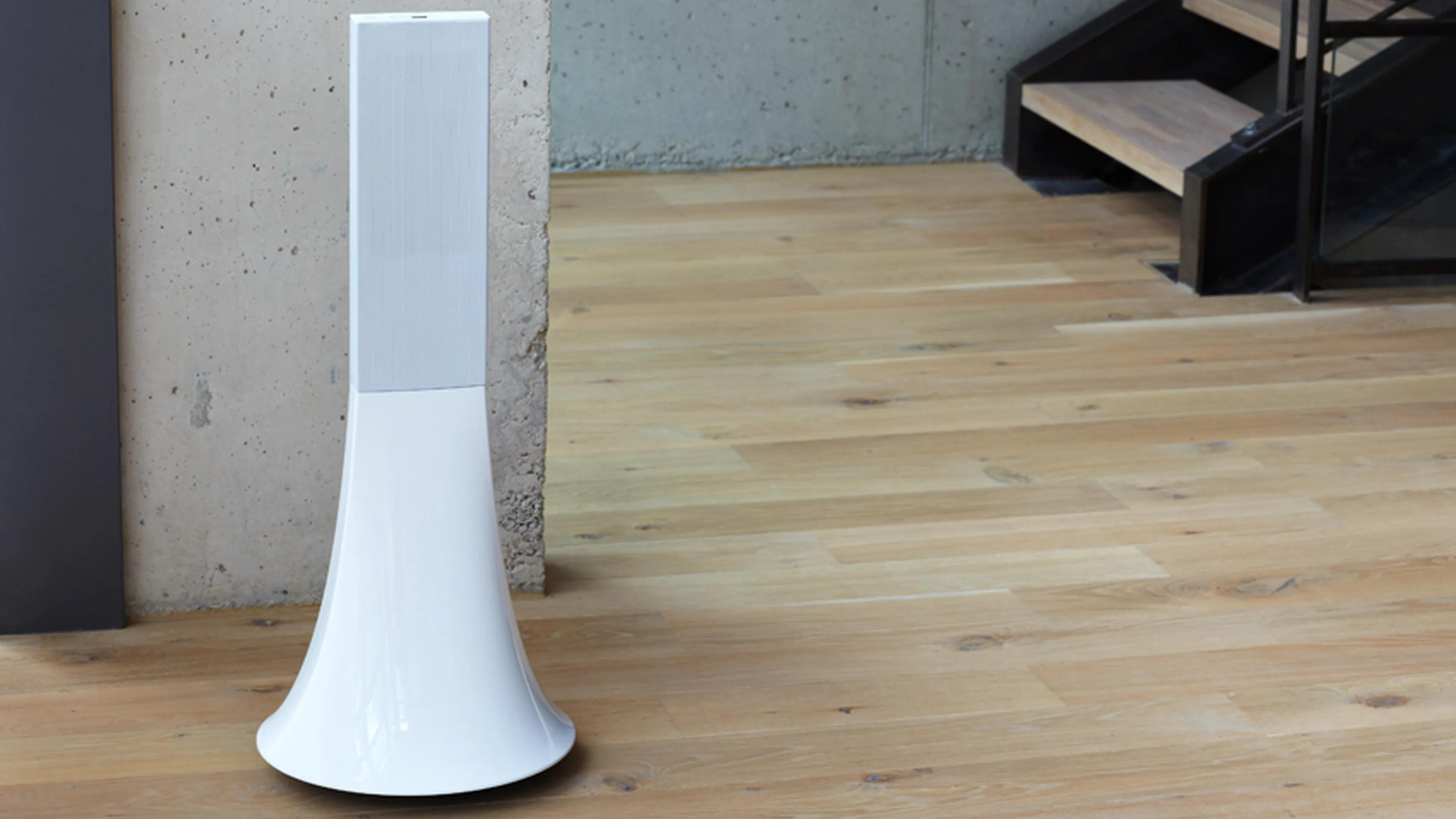 White tower speaker with a flared base standing against a concrete wall on a wood floor by a staircase.
