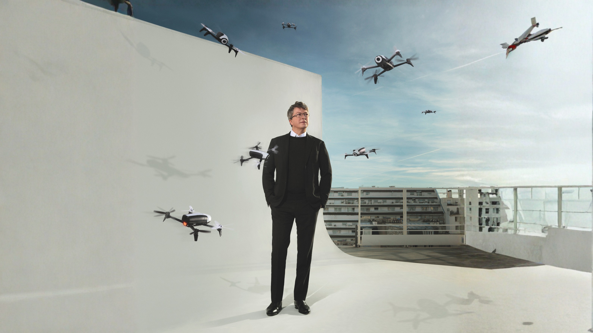 Man in a black suit stands on a white rooftop set surrounded by Parrot quadcopter drones in flight
