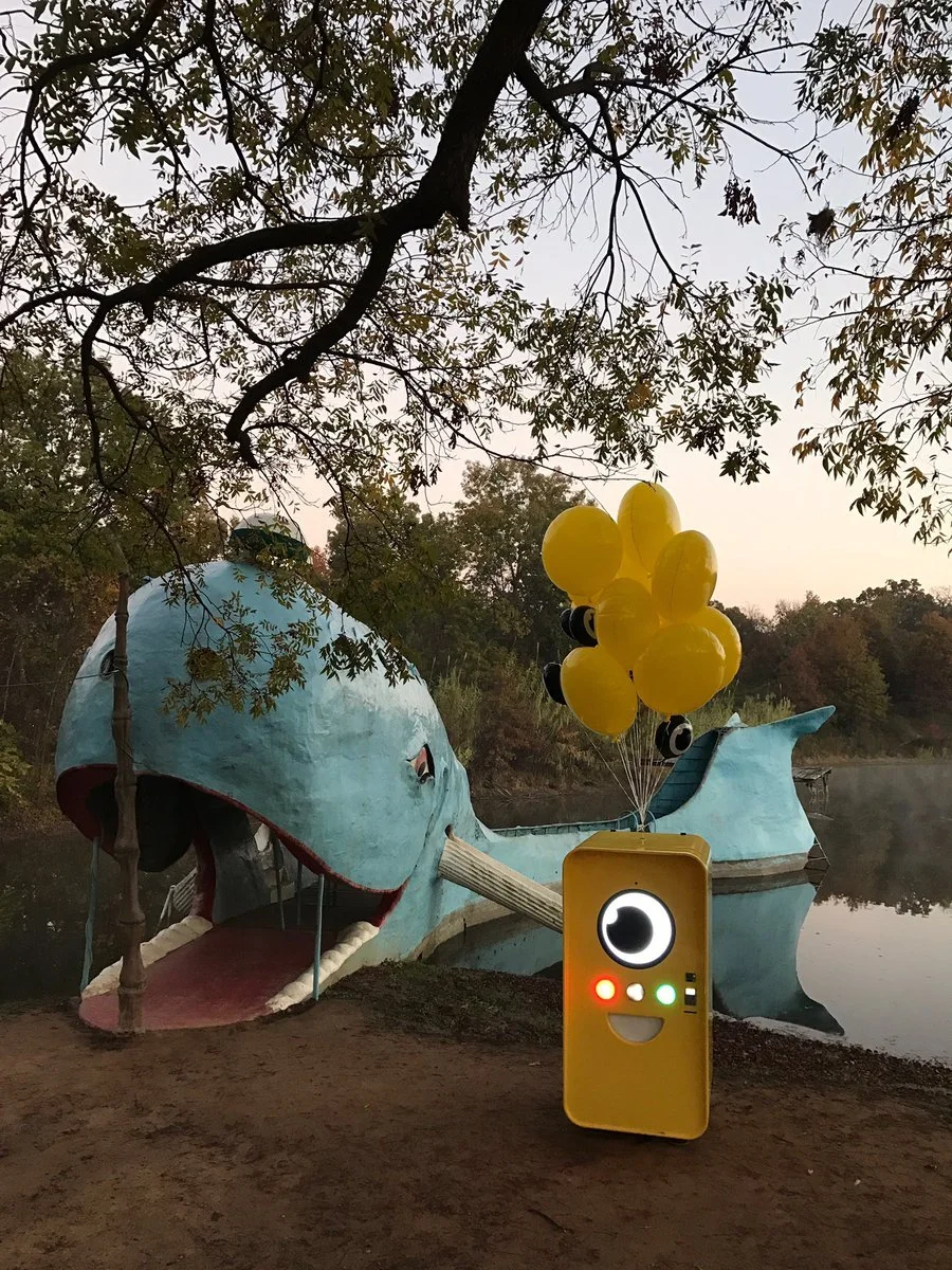 Yellow Snapchat Snapbot with a cartoon eye face and yellow balloons beside the concrete Blue Whale roadside attraction on a lake.