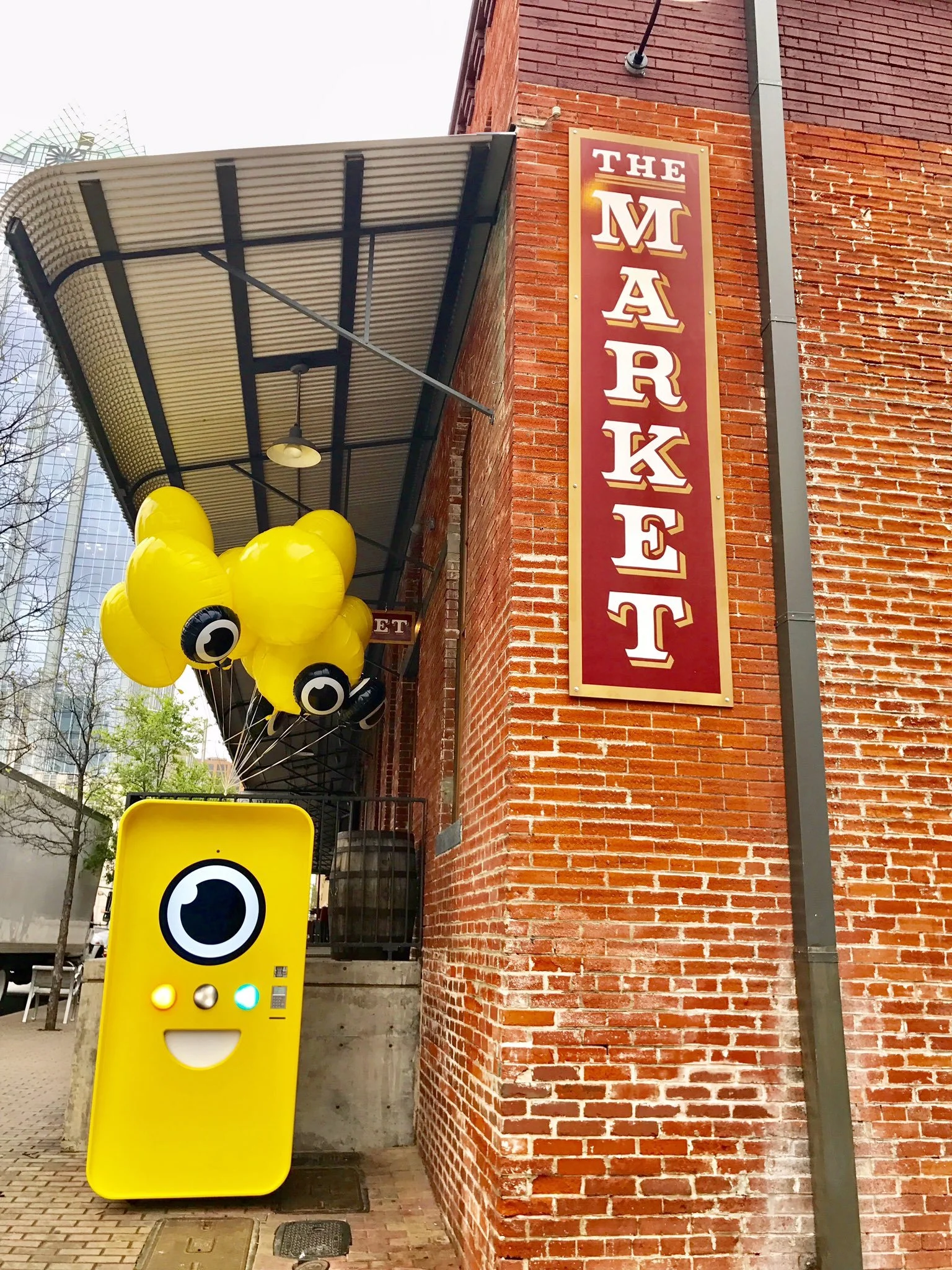 Yellow Snapbot vending machine with balloons beside a brick building and a vertical THE MARKET sign