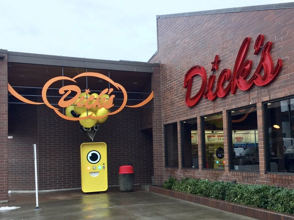 Yellow Snapbot vending machine with balloons outside a brick building under the red Dick's neon sign.