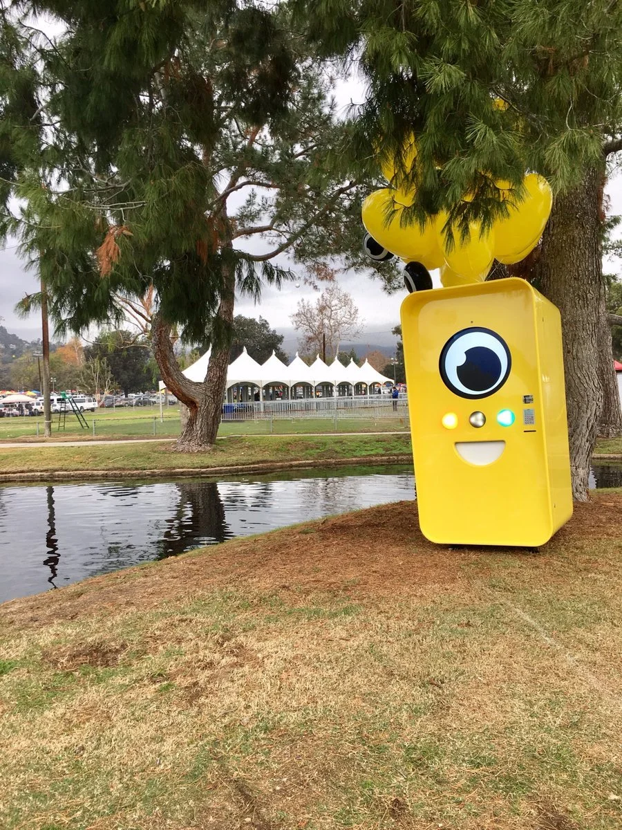 Yellow Snapbot Spectacles vending machine with balloons under a pine tree by a pond, white event tents behind.