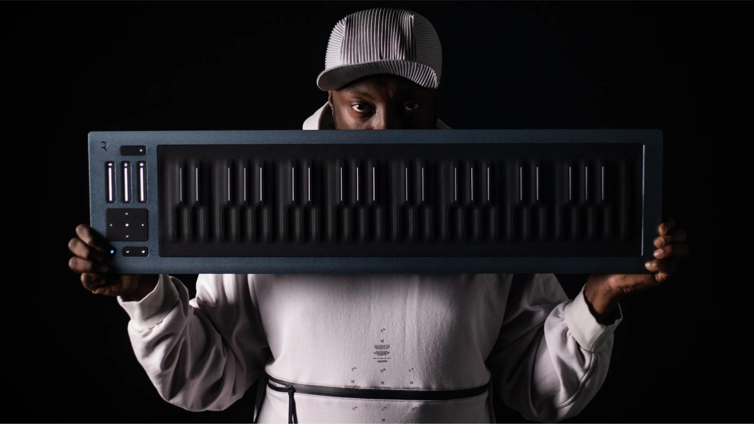 Man in a white hoodie and cap holding a dark ROLI Seaboard keyboard up across his face against a black background