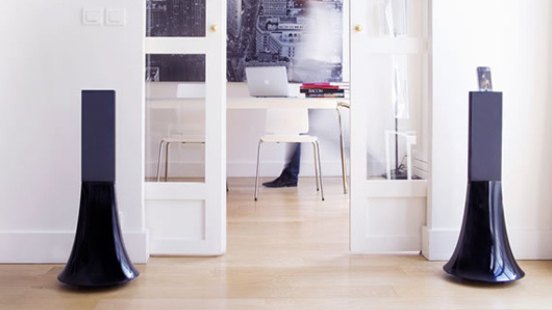 Two black flared tower speakers on a wood floor flanking an open doorway to a bright home office.