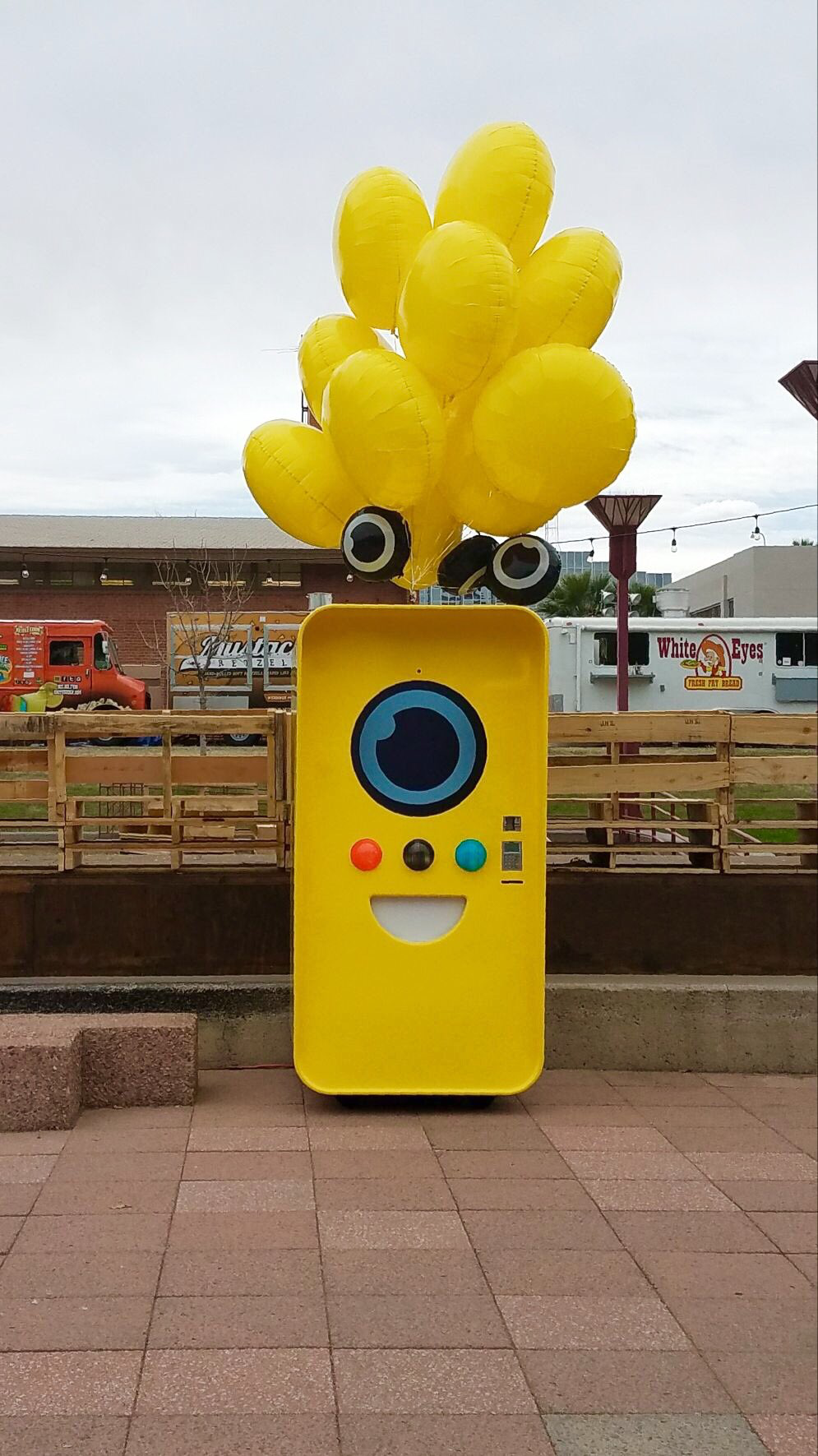 Yellow Snapbot machine with balloons on a plaza, food trucks including White Eyes Fry Bread behind