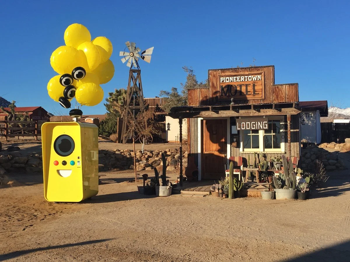 Yellow Snapbot vending machine with balloons in a dirt lot beside the wooden Pioneertown Motel and a windmill.