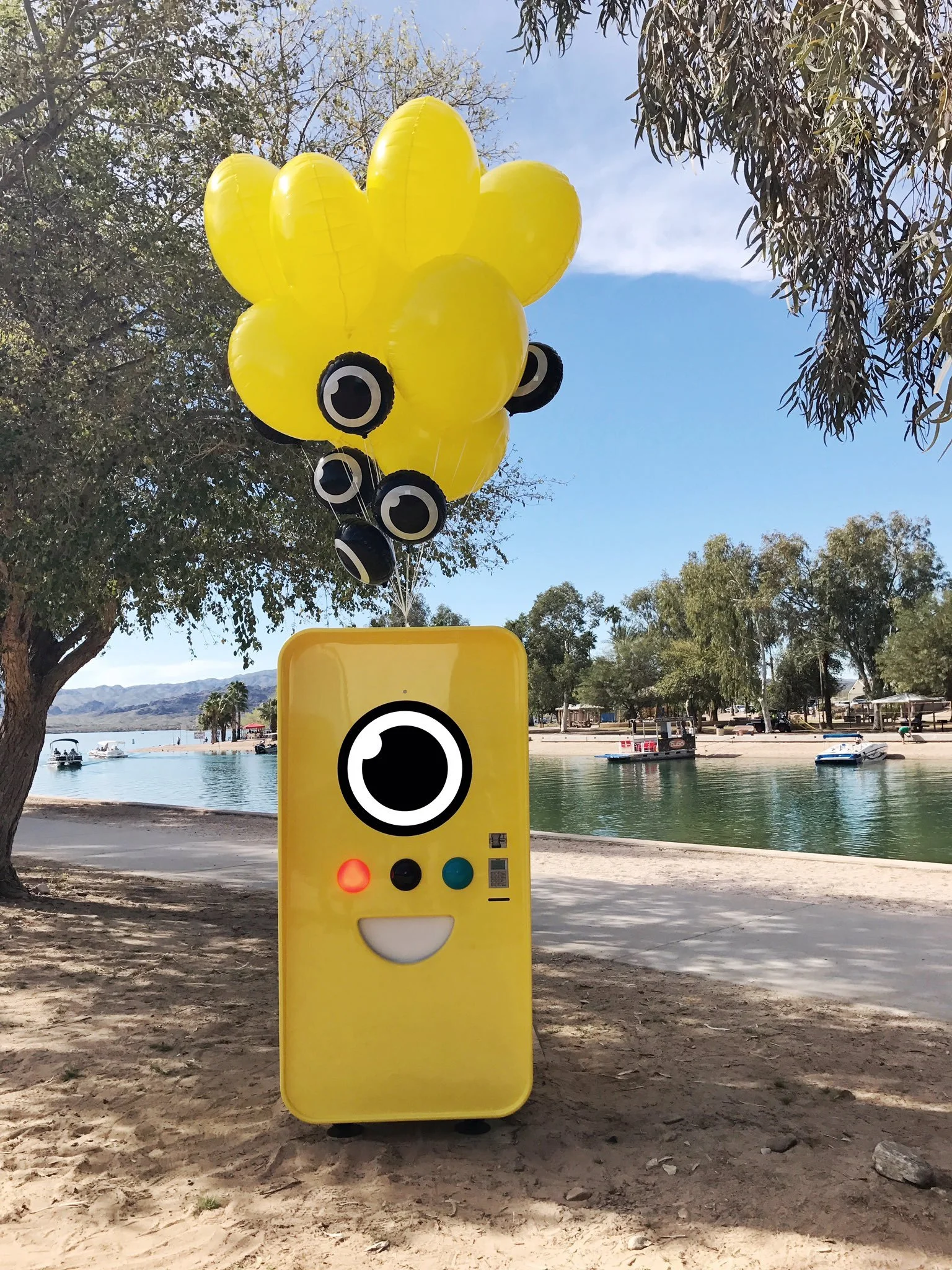 Yellow Snapbot vending machine with a smiling face and yellow balloons on a sandy lakeside beach