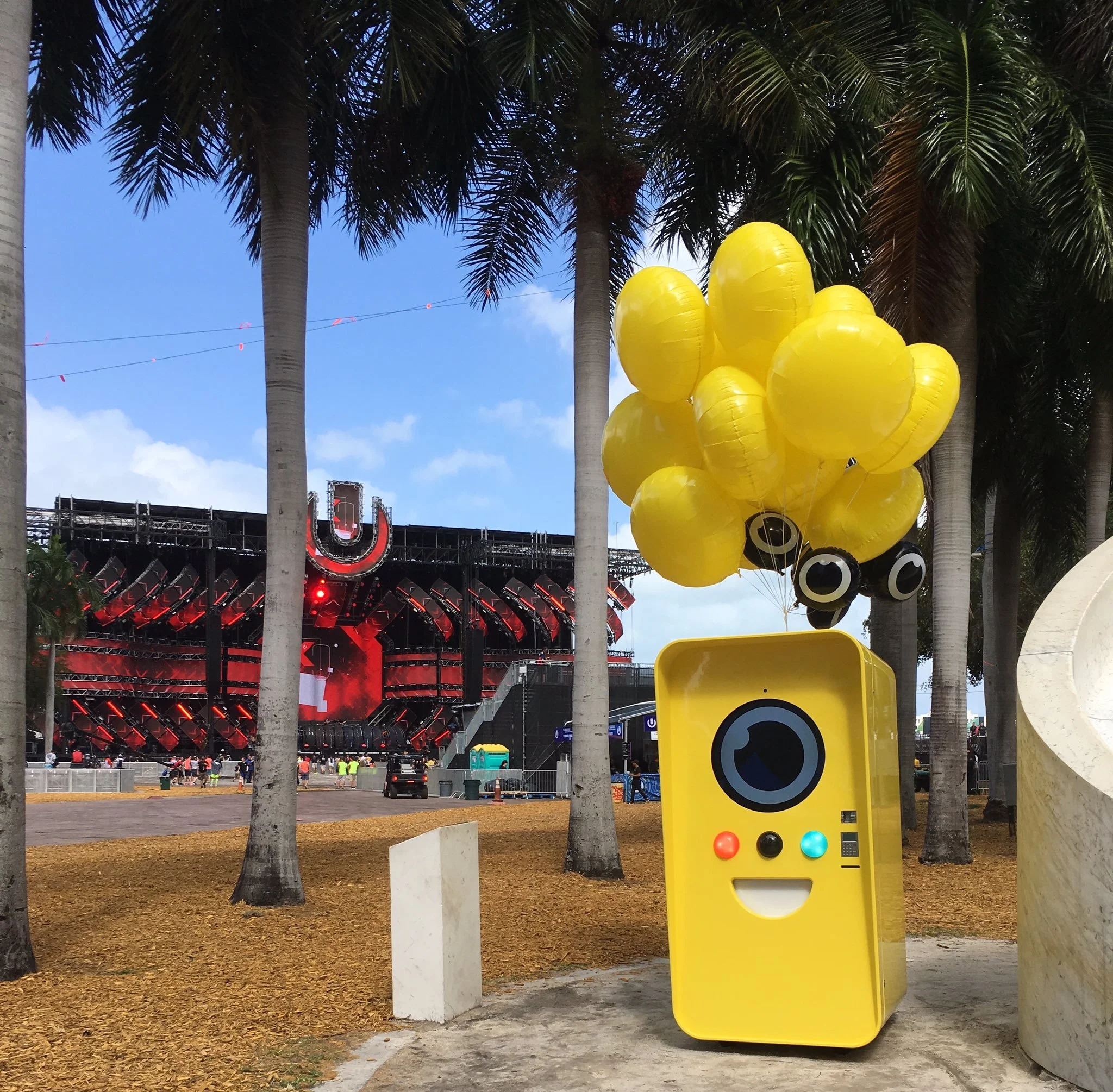 Yellow Snapbot machine with balloons among palm trees, red Ultra Music Festival stage behind