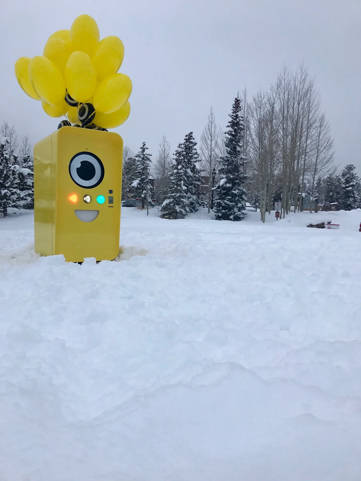 Yellow Snapbot Spectacles vending machine with balloons standing in deep snow among snow-covered evergreens.