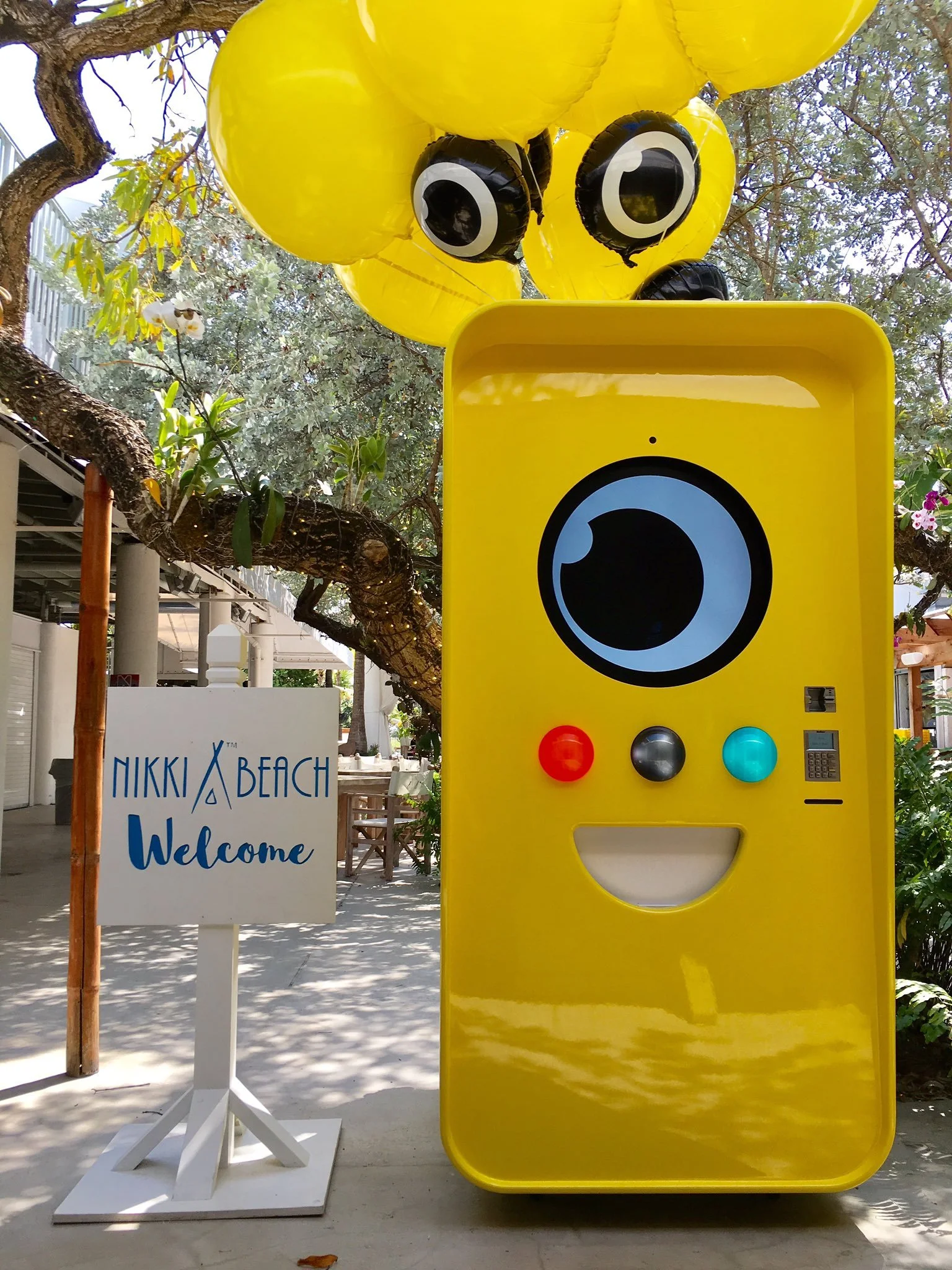 Yellow Snapbot vending robot with camera eye and yellow balloons beside a Nikki Beach Welcome sign