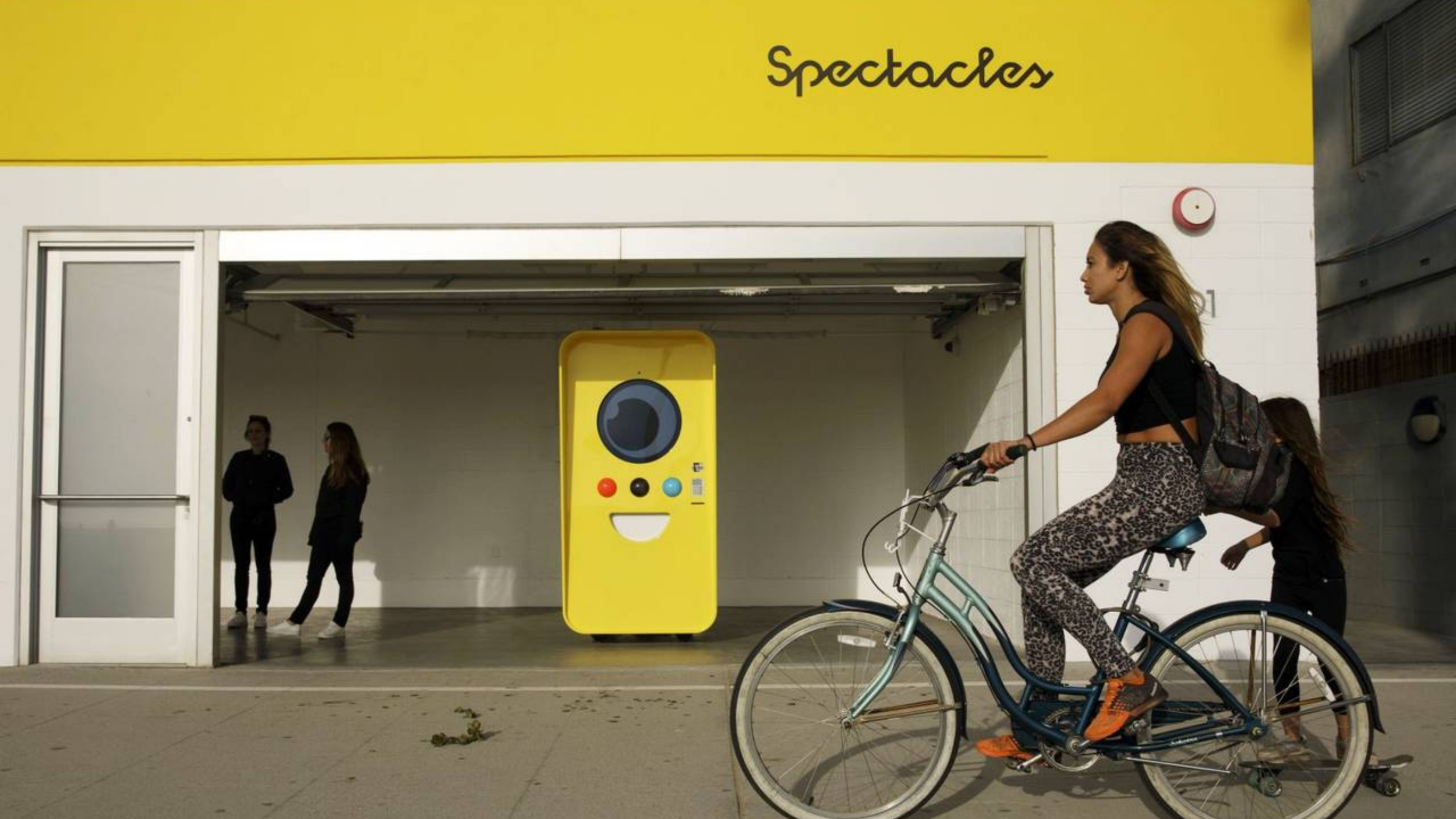 Woman on a bike passes a yellow Snapbot Spectacles vending machine under a 'Spectacles' sign