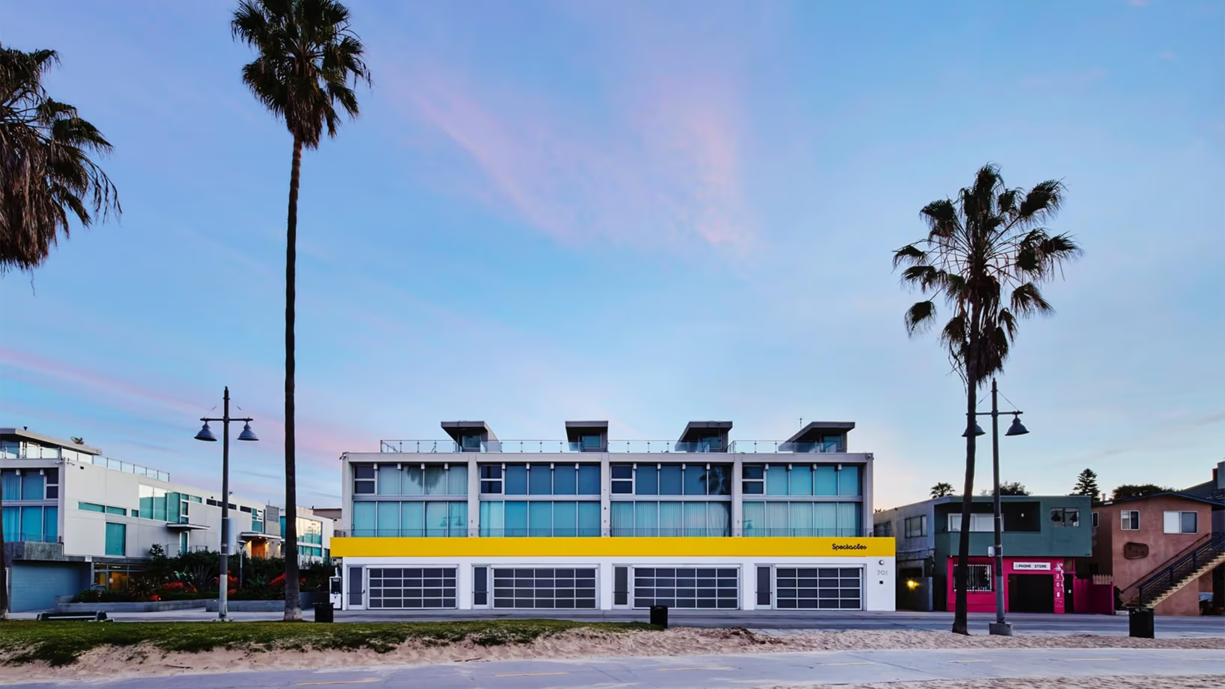Venice Beach building with a yellow 'Spectacles' storefront and garage doors at dusk, framed by palm trees