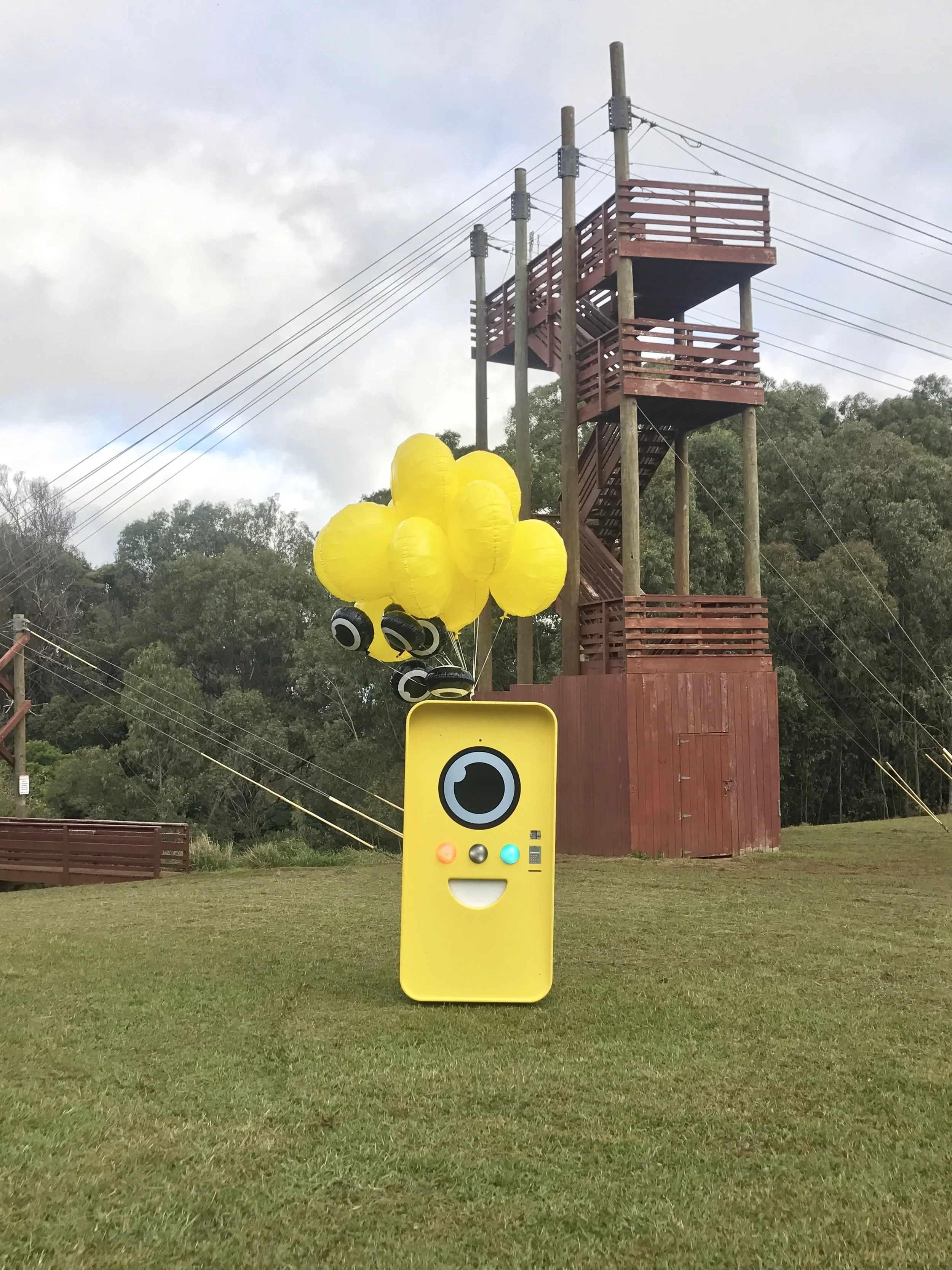 Yellow Snapbot Spectacles vending machine with balloons on a lawn below a tall wooden ropes-course tower.