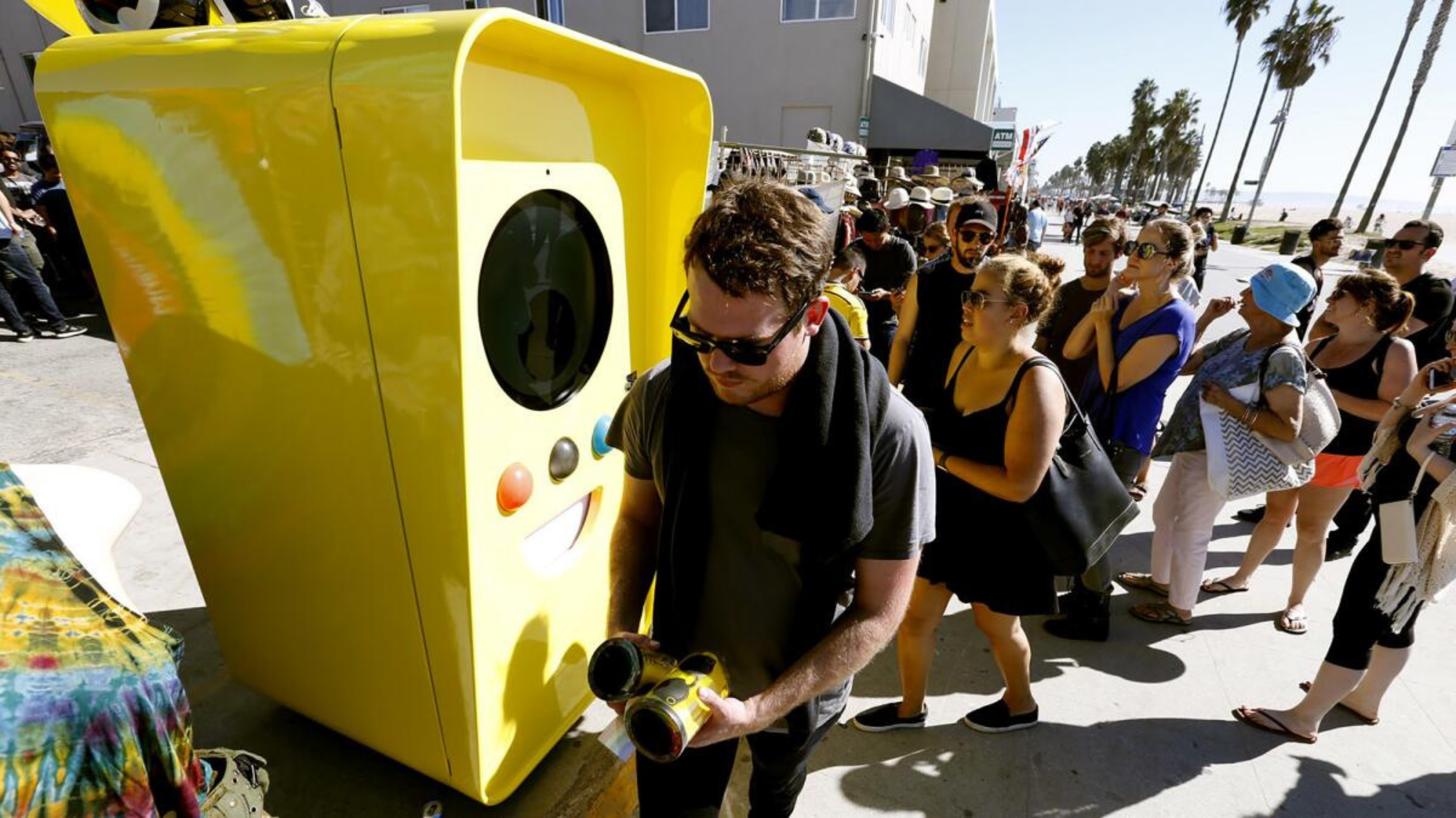 Man in sunglasses holds Spectacles beside a giant yellow Snapbot vending robot as a beachfront crowd lines up