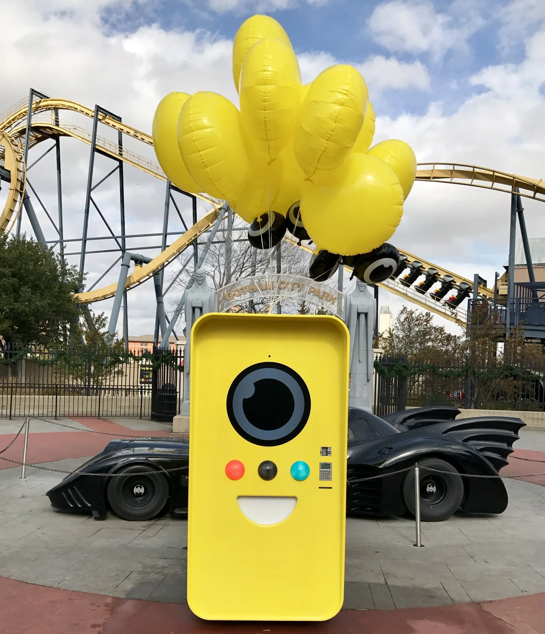 Yellow Snapbot Spectacles vending machine with balloons in front of two black Batmobiles and a Gotham City Park roller coaster.