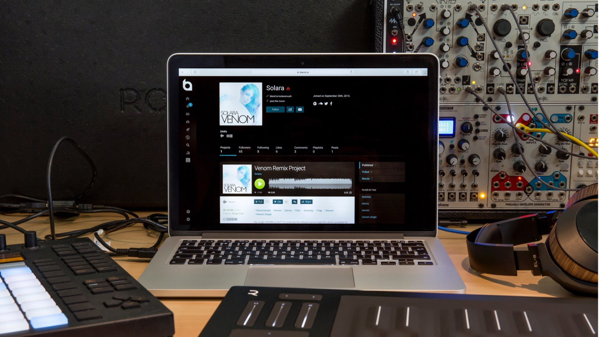 MacBook on a desk showing the blend.io page for artist Solara's Venom Remix Project, flanked by a modular synth, MIDI pads and headphones