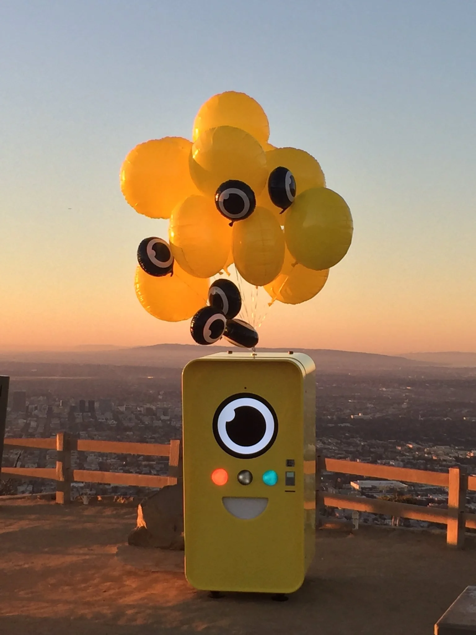 Yellow Snapbot Spectacles vending machine with balloons on a hilltop overlook above the Los Angeles skyline at sunset.