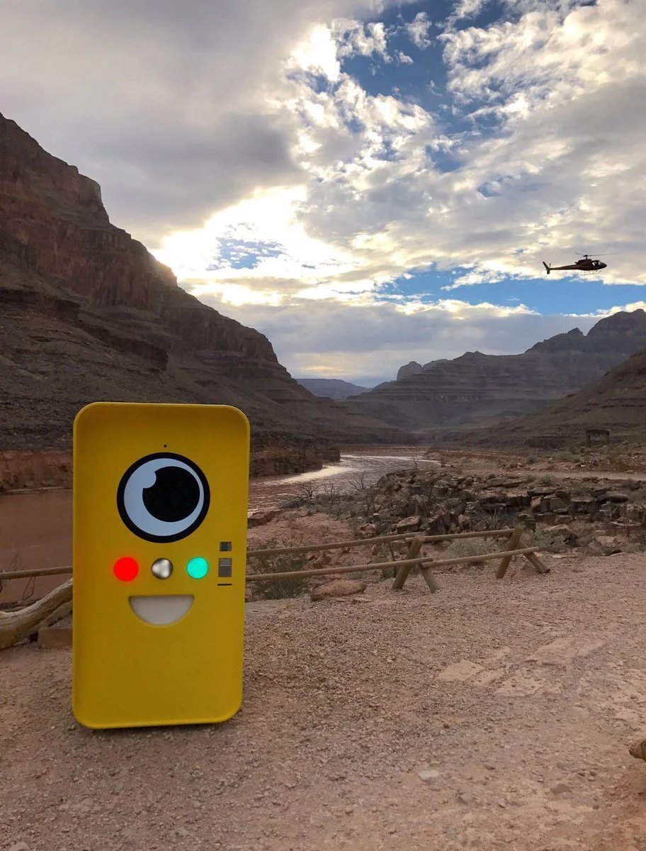 Yellow Snapbot Spectacles vending machine on a canyon riverbank, with a helicopter flying over the Grand Canyon behind it.