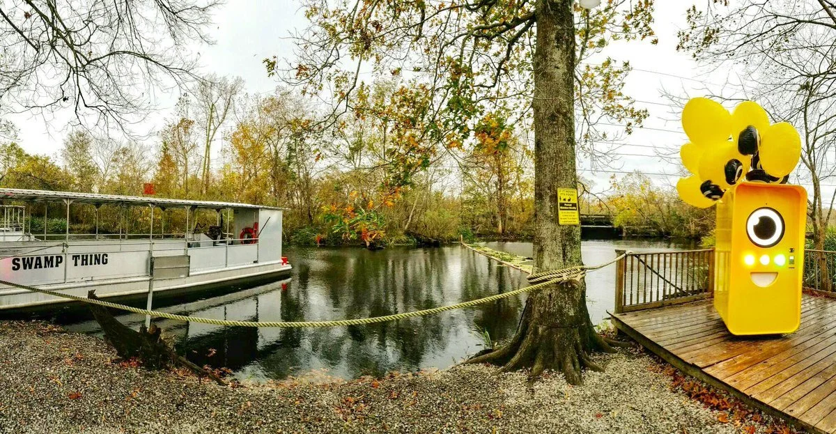Yellow Snapbot Spectacles vending machine on a wooden dock by a swamp, beside a Swamp Thing tour boat.