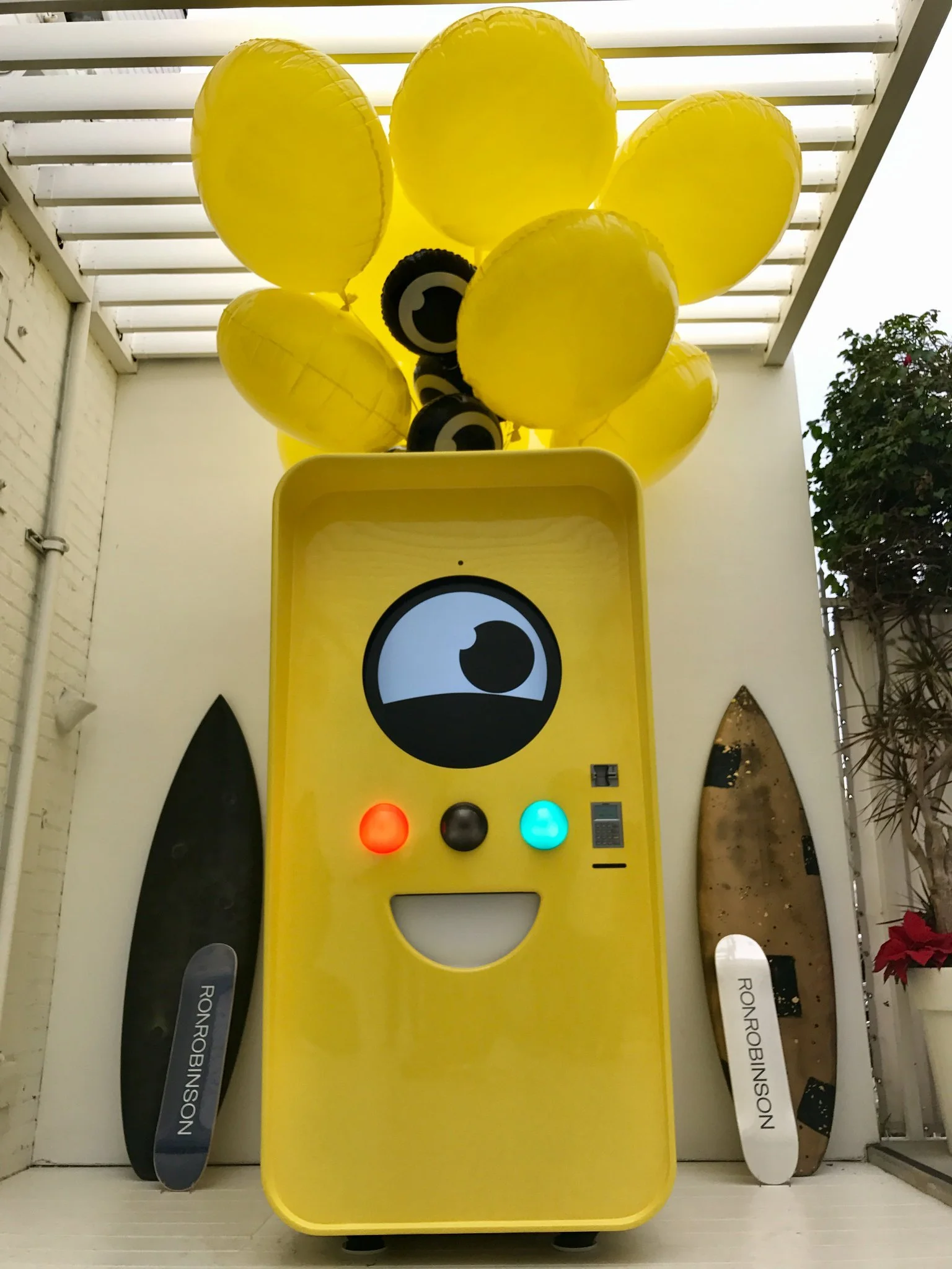 Yellow Snapbot Spectacles vending machine with cartoon eye face and yellow balloons, between two Ron Robinson surfboards.
