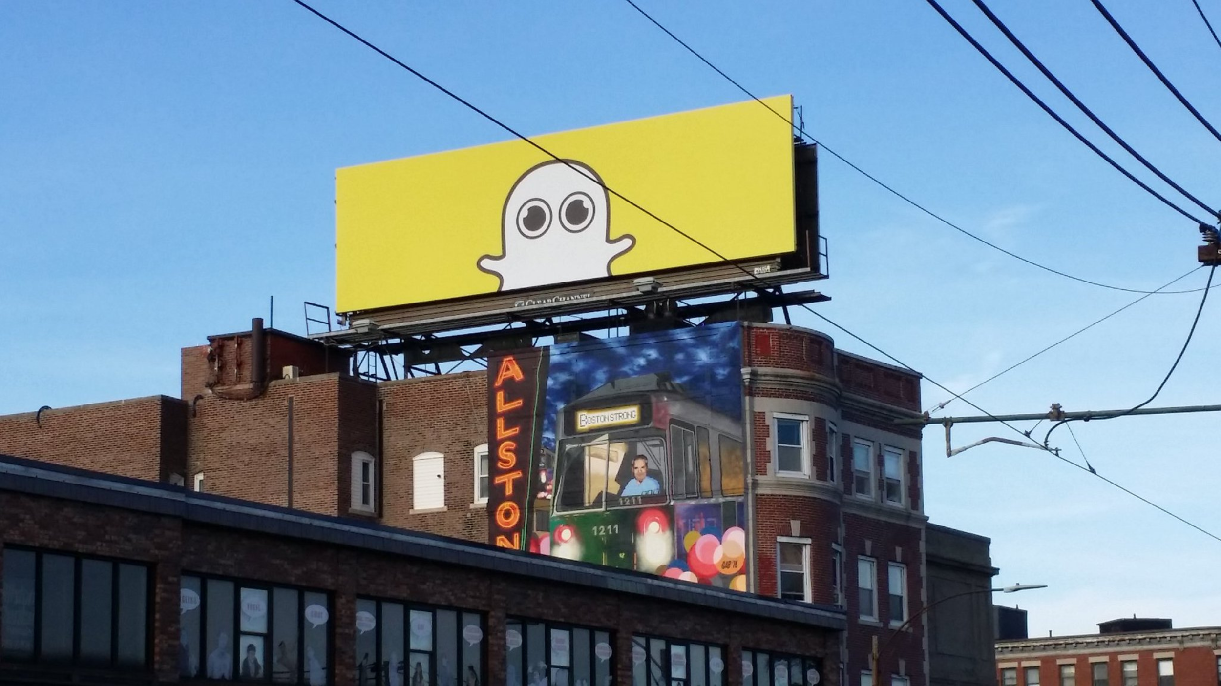 Yellow Snapchat ghost billboard atop an Allston brick building against blue sky